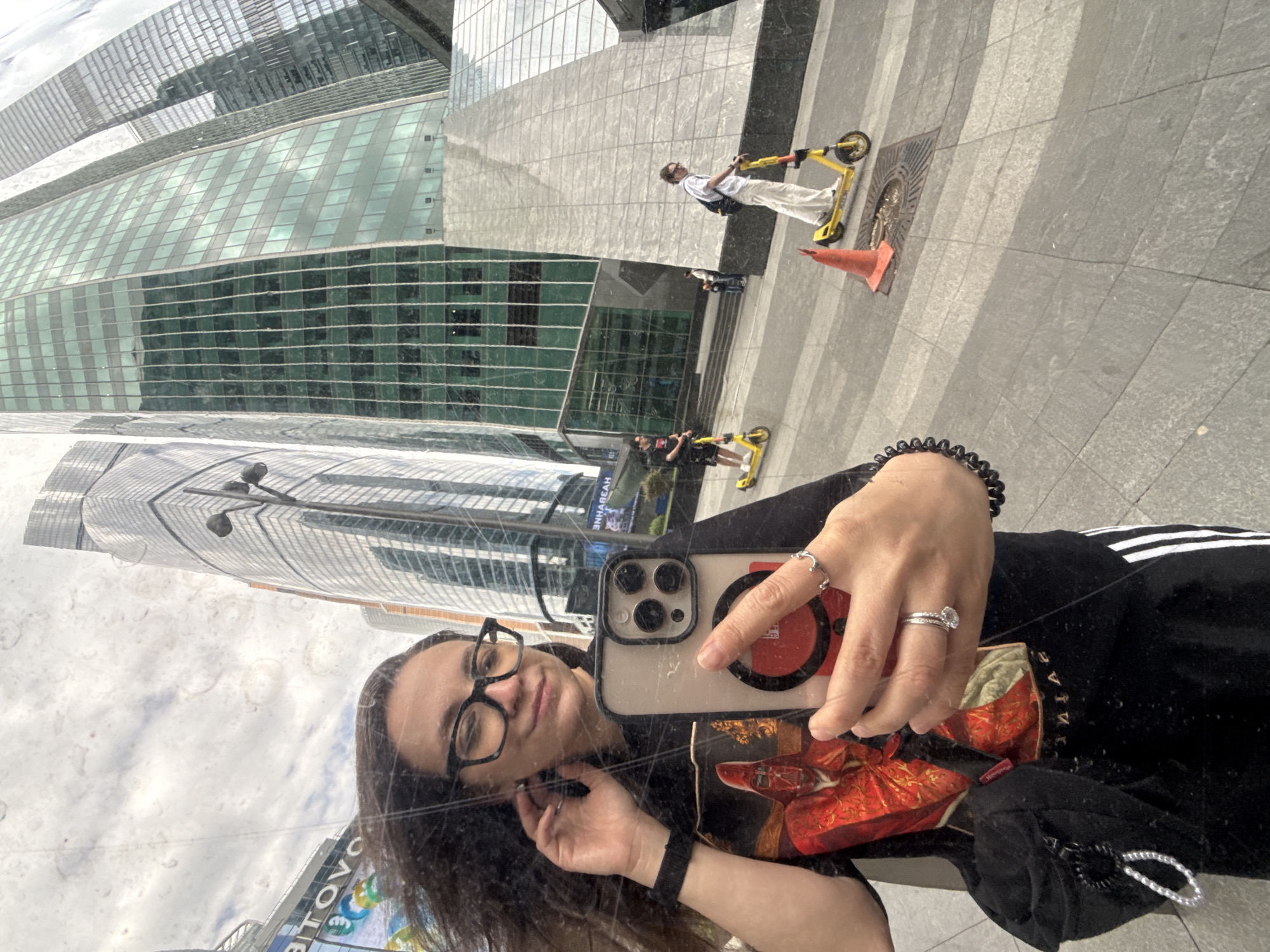 A reflection selfie shows a person with brown hair and glasses holding a smartphone with a red phone grip. The reflection reveals city architecture, including glass skyscrapers. In the background, people ride scooters near an orange traffic cone.