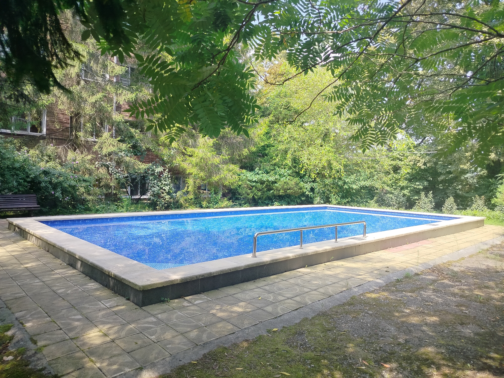 The image depicts an outdoor swimming pool with a blue tiled bottom, surrounded by a stone deck. A metal handrail is installed along one side of the pool. In the background, lush green trees and foliage are visible, with a brick building partially obscured by vegetation. A wooden bench can be seen to the left of the pool. The scene appears to be set in a residential or recreational area, possibly a garden or backyard. The lighting suggests it is daytime, with sunlight filtering through the trees. There are no people visible in the image. The location, Krasnyi Most, Russia, is not discernible from the visual elements of the photograph.