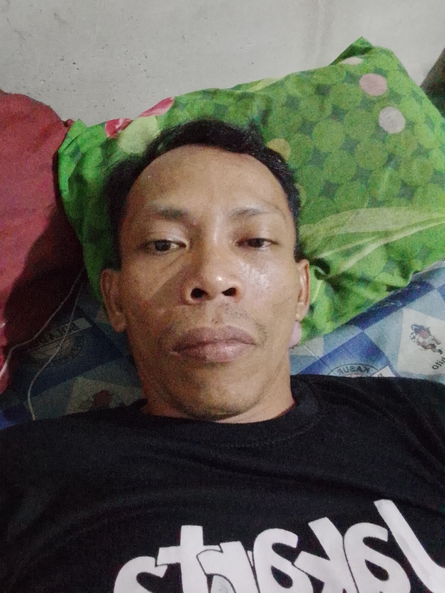 A man with dark hair and skin lies on a bed in Bukit Ketok, Indonesia. He wears a black shirt with white writing. A green and pink patterned pillow rests behind his head, and a red pillow is to his left. The bedsheets are blue and white.