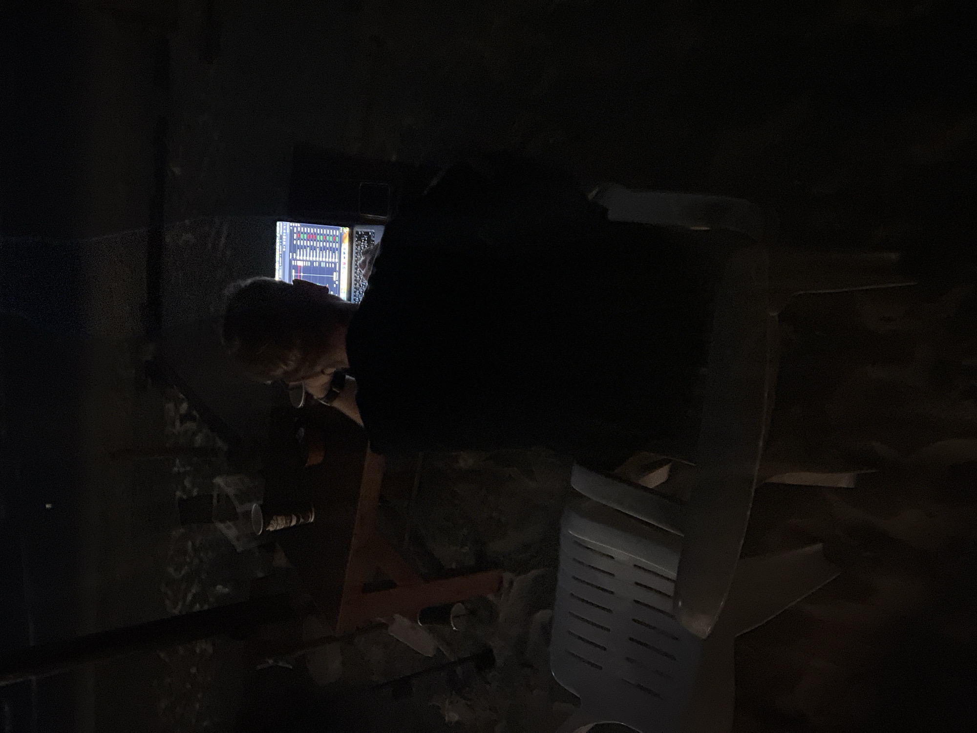 This media file shows a person hunched over a desk in a dimly lit environment, likely a home studio or office. The person appears to be focused on a computer monitor, which displays a visual representation of audio waveforms, suggesting they are involved in audio production or editing. To the right of the monitor is what looks like a mixing board or audio interface, further supporting the idea of audio work. The background is dark and somewhat cluttered, with glimpses of what could be soundproofing material or acoustic treatment on the walls. The overall atmosphere is one of concentration and creative work, as the subject is deeply engrossed in their task. The lighting is minimal, primarily coming from the screen itself, which casts a soft glow on the person's face and hands. This lighting choice emphasizes the focus on the technology and the work being done. The image's orientation is rotated, suggesting it was taken with the camera held sideways.