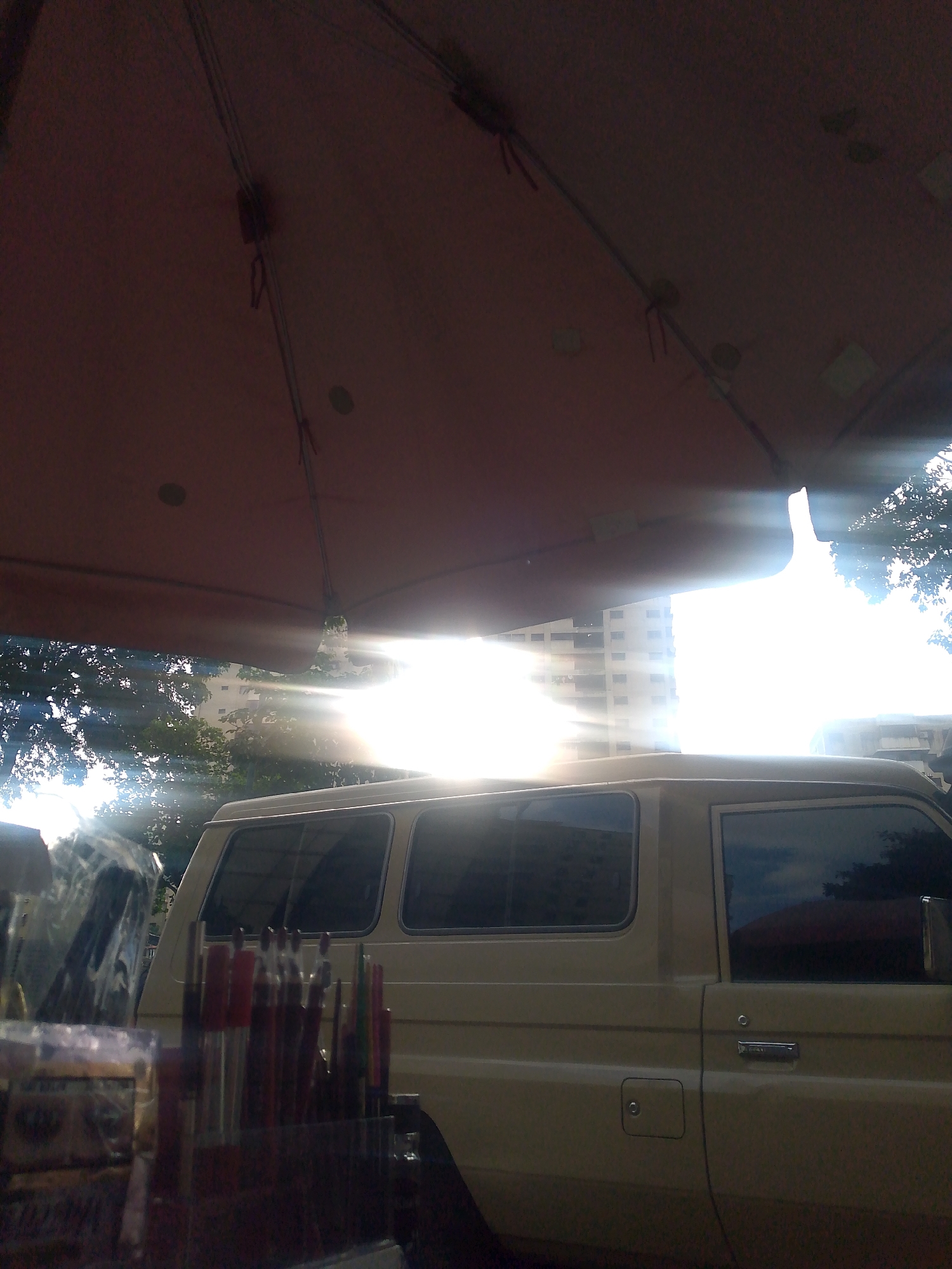 The image captures an outdoor scene in Caracas, Venezuela, with a large umbrella obscuring the top portion of the frame, casting a shadow over much of the foreground. Bright sunlight beams through the umbrella, creating lens flare and highlighting the environment. In the mid-ground, a beige van is parked. The van has tinted windows and appears to be a utility or transport vehicle. In the immediate foreground, on the left side of the image, a display of items, possibly art supplies or small crafts, is visible. These items are colorful and appear to be arranged in a holder. Trees with lush green foliage are visible behind the van, and in the distance, a tall, multi-story building stands, suggesting an urban setting. The overall impression is a market stall or an outdoor vendor area, possibly with the van used for transport or as part of the setup. The lighting conditions suggest it is daytime, likely mid-morning or afternoon, given the angle of the sun.
