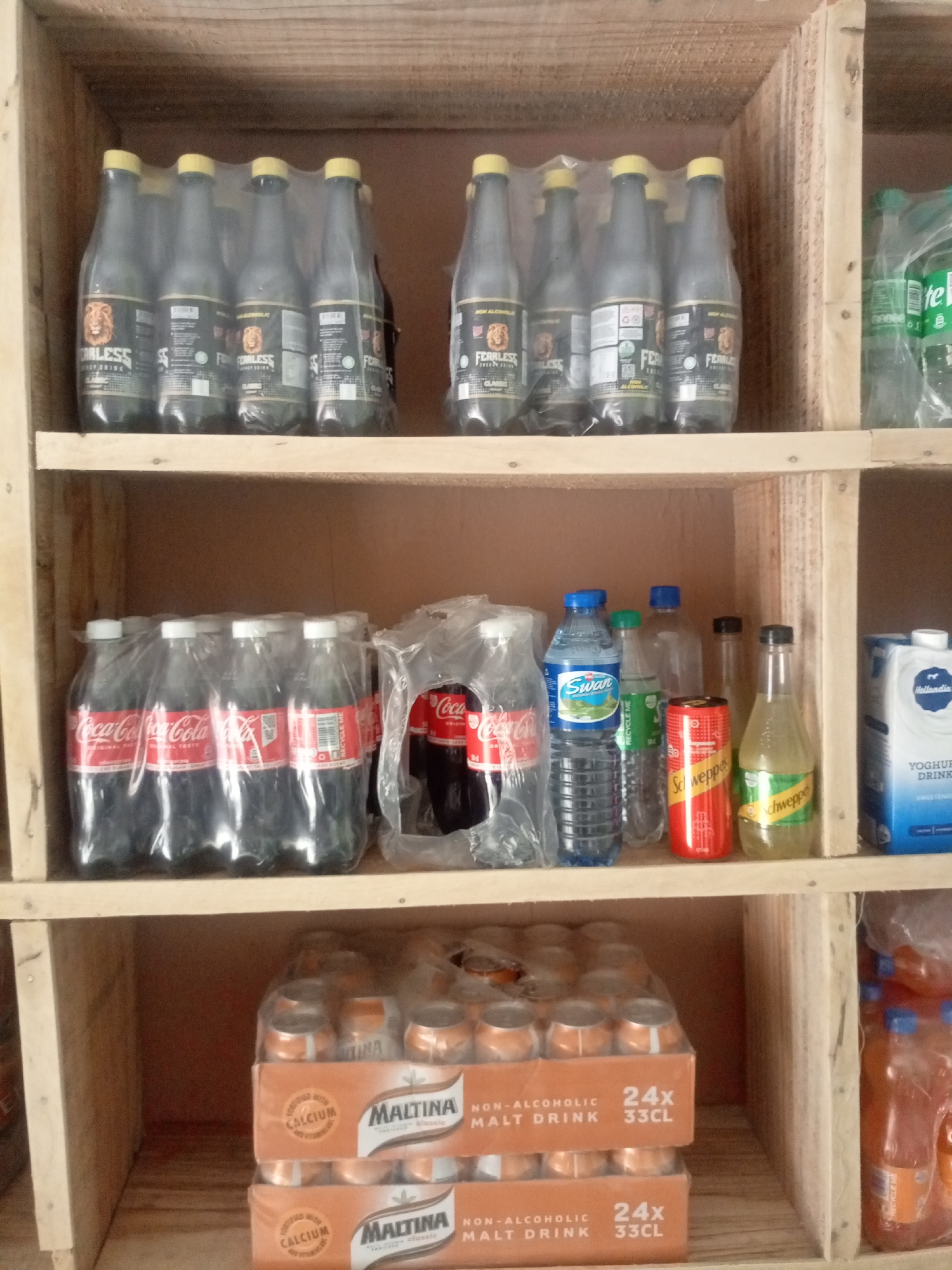 The image depicts a wooden shelf filled with various beverages, likely in a retail setting in Jos, Nigeria. On the top shelf, several bottles of "FEARLESS" energy drink are stacked, some in plastic wrap. To the right, a few bottles of Sprite are visible. The middle shelf holds a multipack of Coca-Cola bottles, followed by a can of Schweppes and another bottle of Schweppes ginger ale. A carton of Hollandia Yoghurt Drink is on the far right. The bottom shelf features two stacked cartons of "MALTINA" non-alcoholic malt drink. The overall environment appears to be a storage area or a small shop.