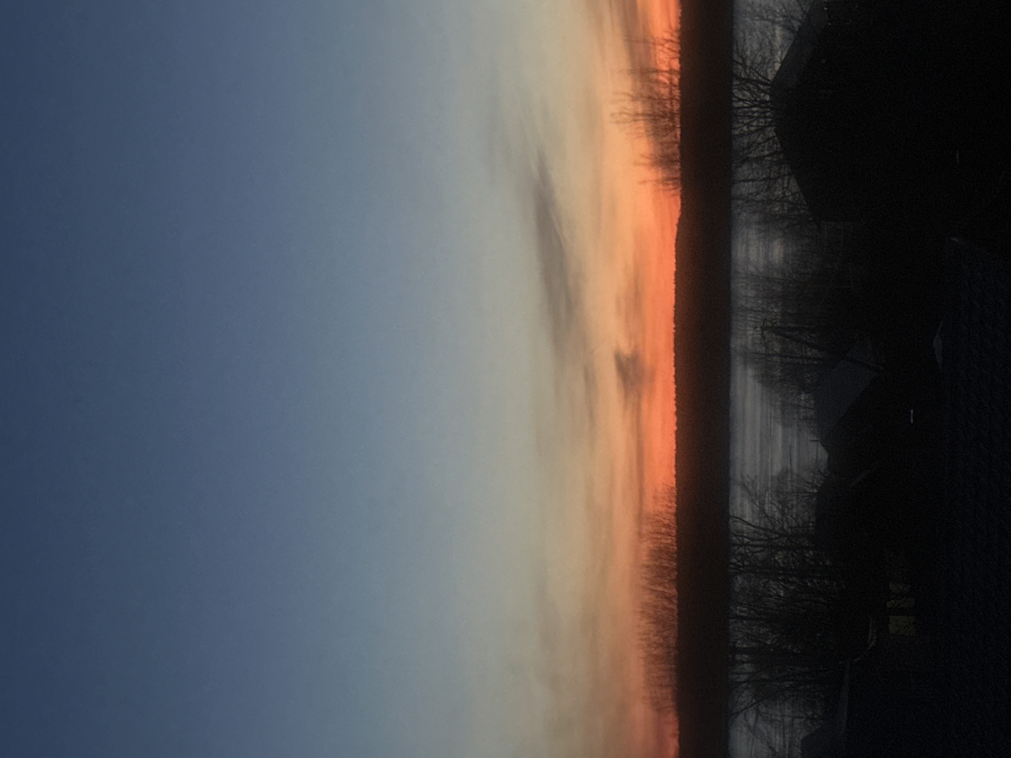The image shows a dusk or dawn scene in what appears to be a rural area. The sky is a gradient of blue at the top transitioning to orange and red near the horizon, suggesting either sunset or sunrise. Along the horizon line, there is a line of trees, or possibly a forest edge, in silhouette. Adjacent to the trees is a body of water, which is reflecting the colors of the sky. In the foreground, a dark, indistinct structure or vegetation is visible. Specific details about the city of Chaikovsky, Russia cannot be verified from the provided image. There are no people visible in the image.