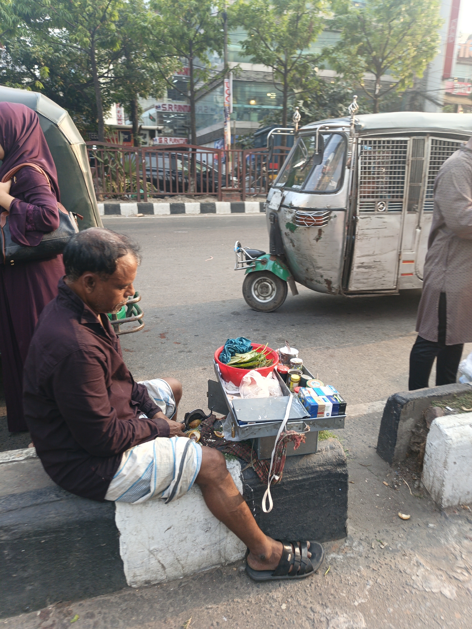 This outdoor, eye-level photograph captures a vibrant street scene in Dhaka, Bangladesh, on a bright, sunny day. The primary subject is a middle-aged man, with receding dark hair and a short beard, wearing a dark maroon long-sleeved shirt and a white and blue striped lungi-style garment. He is seated on a low concrete curb, looking down intently at a small, portable vendor's tray placed on a dark block beside him. He wears black sandals on his feet.
The vendor's tray is laden with various items for sale. A red plastic bowl holds green betel leaves, and several small containers, likely holding spices, nuts, or tobacco for paan, are arranged on the tray. Packs of cigarettes, including visible brands like "STAR" and "BENSON & HEDGES", are also part of his merchandise. Next to the tray, on a patterned cloth, are broken coconut shells or similar husks.
To the left of the man, partially visible, stands a woman wearing a dark maroon hijab and a matching long-sleeved dress, carrying a black shoulder bag. Behind the man, a distinctive three-wheeled auto-rickshaw, commonly known as a CNG auto in Bangladesh, is parked by the roadside. It has a metallic grey, weathered body with a prominent front grille and a caged passenger compartment. Another person, dressed in a brown-grey long tunic, is partially visible to the right of the auto-rickshaw.
The background reveals a bustling urban environment. A multi-lane road with a black and white striped pedestrian crossing is visible. A metal fence separates the sidewalk from a commercial area with several green trees. Beyond the trees, modern buildings with glass facades line the street. Visible text on one of the buildings clearly reads "SRINI FURNITURE" and "SRINI F...N", indicating a furniture store. The scene portrays a typical moment of roadside vending in a busy city.