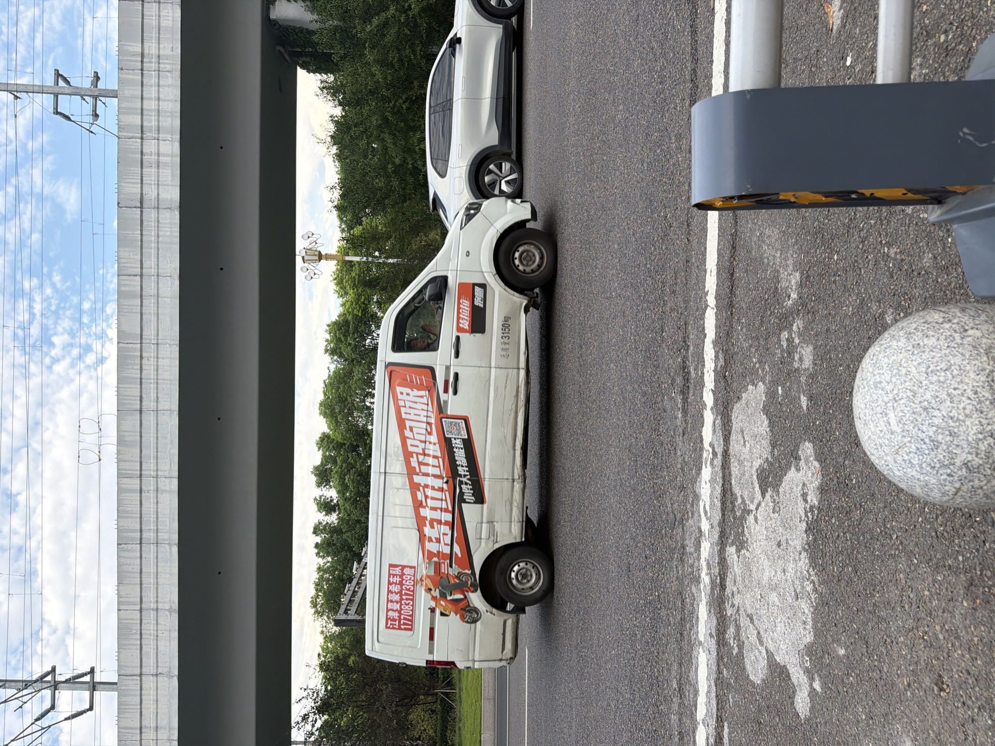 A white delivery van with red and black decals is driving on a road. The van has Chinese text on it, including "江津豪泰车队" (Jiangjin Haotai Team) and a phone number "17708317369". A white SUV is driving in the lane next to the van. The road is lined with trees and a pedestrian walkway. Overhead, a concrete bridge and power lines are visible against a cloudy sky. The scene appears to be in Jiangjin, China.