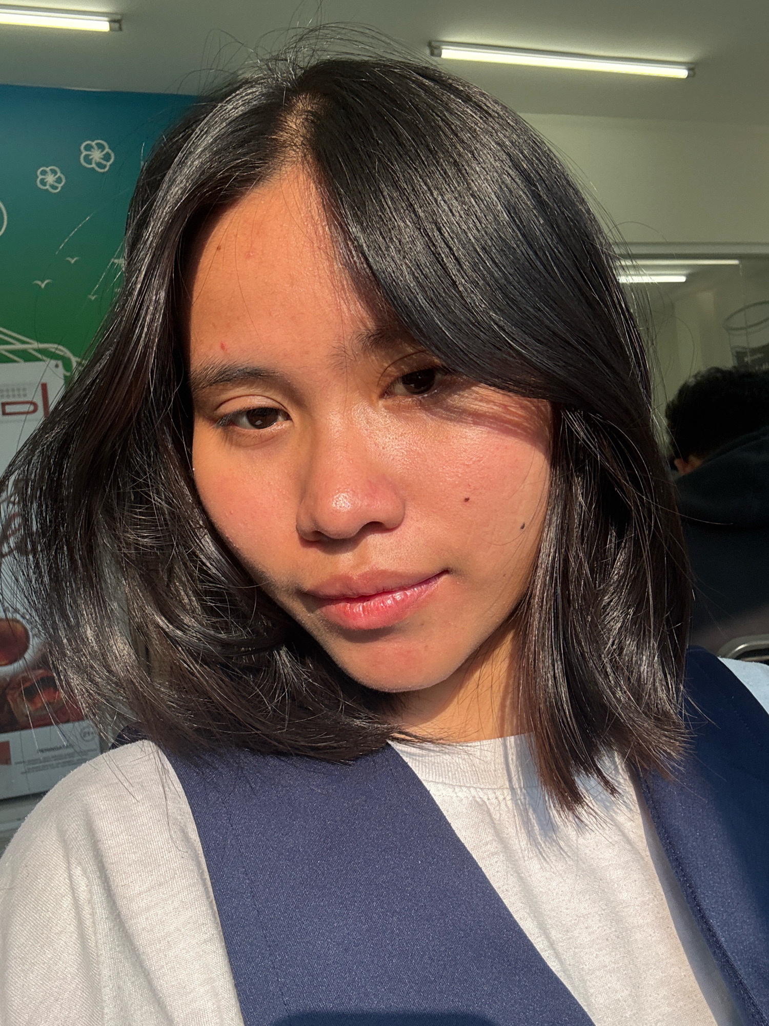 The image is a close-up selfie of a young woman with dark hair and fair skin, taken outdoors in East Jakarta, Indonesia. She is wearing a white t-shirt under a dark blue vest. The background includes a wall with a mural and fluorescent lighting. The sunlight casts a shadow across her face.