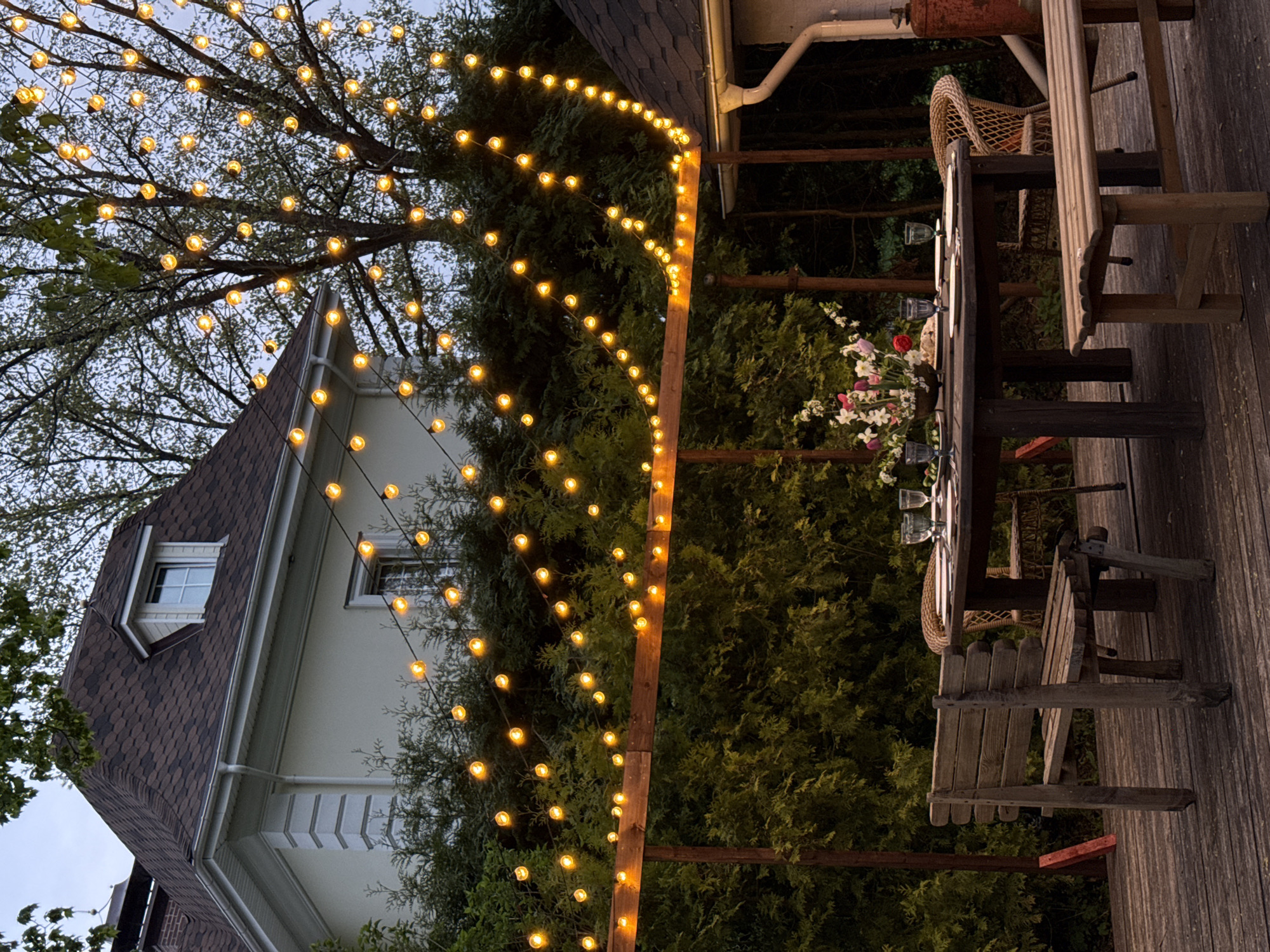 The image depicts an outdoor dining setting. A long wooden table is set for a meal, with plates, glasses, and a floral arrangement in the center. Around the table are wooden chairs and a wicker chair. Overhead, string lights are hung, creating a warm glow. The lights are strung between trees, a wooden pergola, and the side of a house. The house has a dark roof, white siding, and a window. The setting is surrounded by green foliage, suggesting a garden or wooded area. The image was not taken in Sapronovo, Russia.