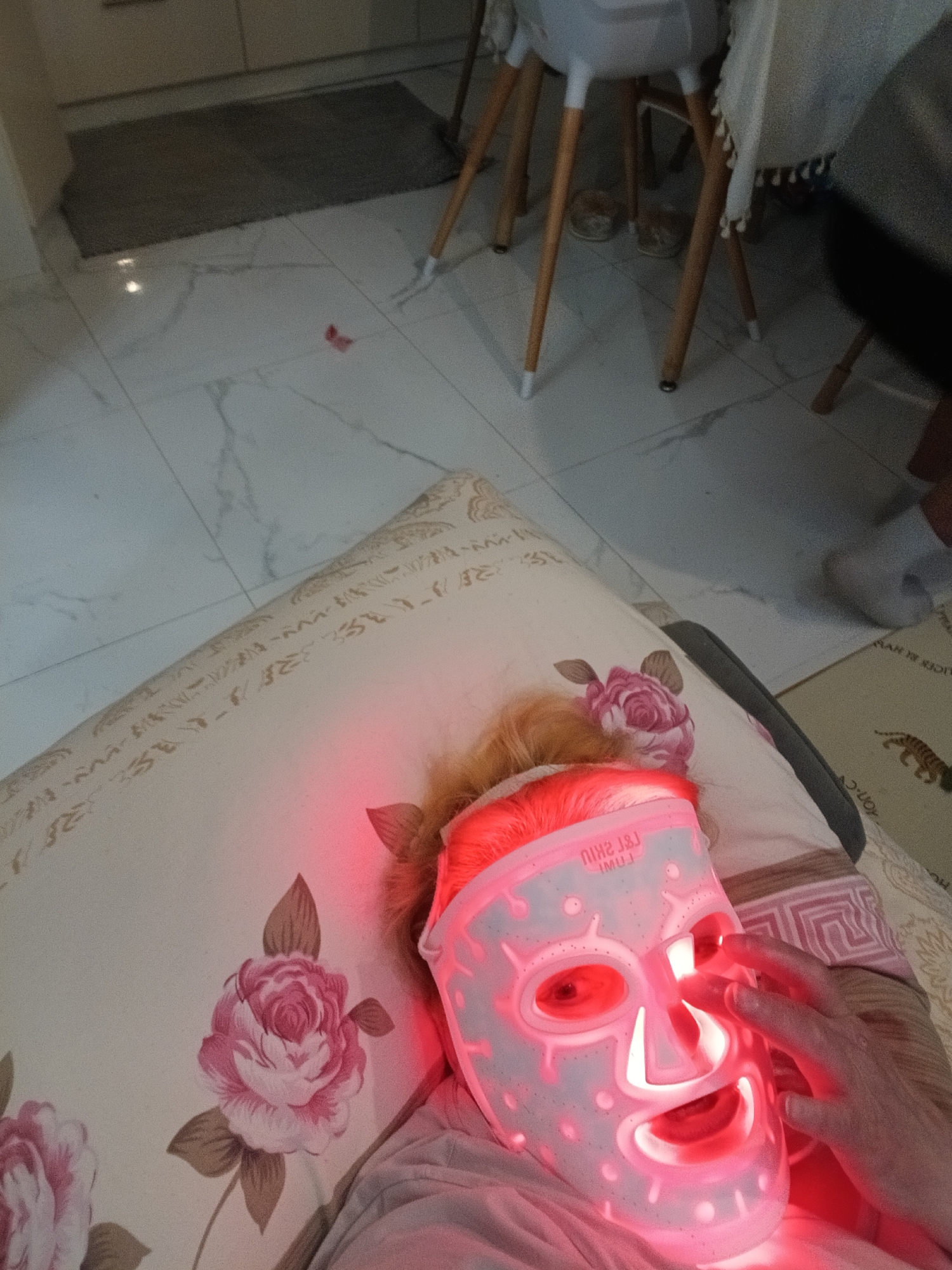 An individual is depicted in an indoor residential setting, likely in the city of Odintsovo, Russia, lying on a bed or couch. The person's head rests on a pillow with a pink rose floral pattern. They are wearing a white or translucent LED facial mask, labeled "L&L SHIU" (reversed), which emits intense red light across their face and light-colored hair. One hand is positioned near the mask, fingers extended towards the eye area. The floor is composed of white tiles with gray veining. In the background, a dark gray rectangular mat is visible on the floor, along with the lower portions of a white high chair with wooden legs and another piece of furniture draped with a light-colored fabric. A small red object lies on the tiled floor. To the right, a foot wearing a light-colored slipper or sock is visible on a beige rug featuring a tiger print and partially visible text. The specific city of Odintsovo, Russia, is not identifiable from visual elements within the image.