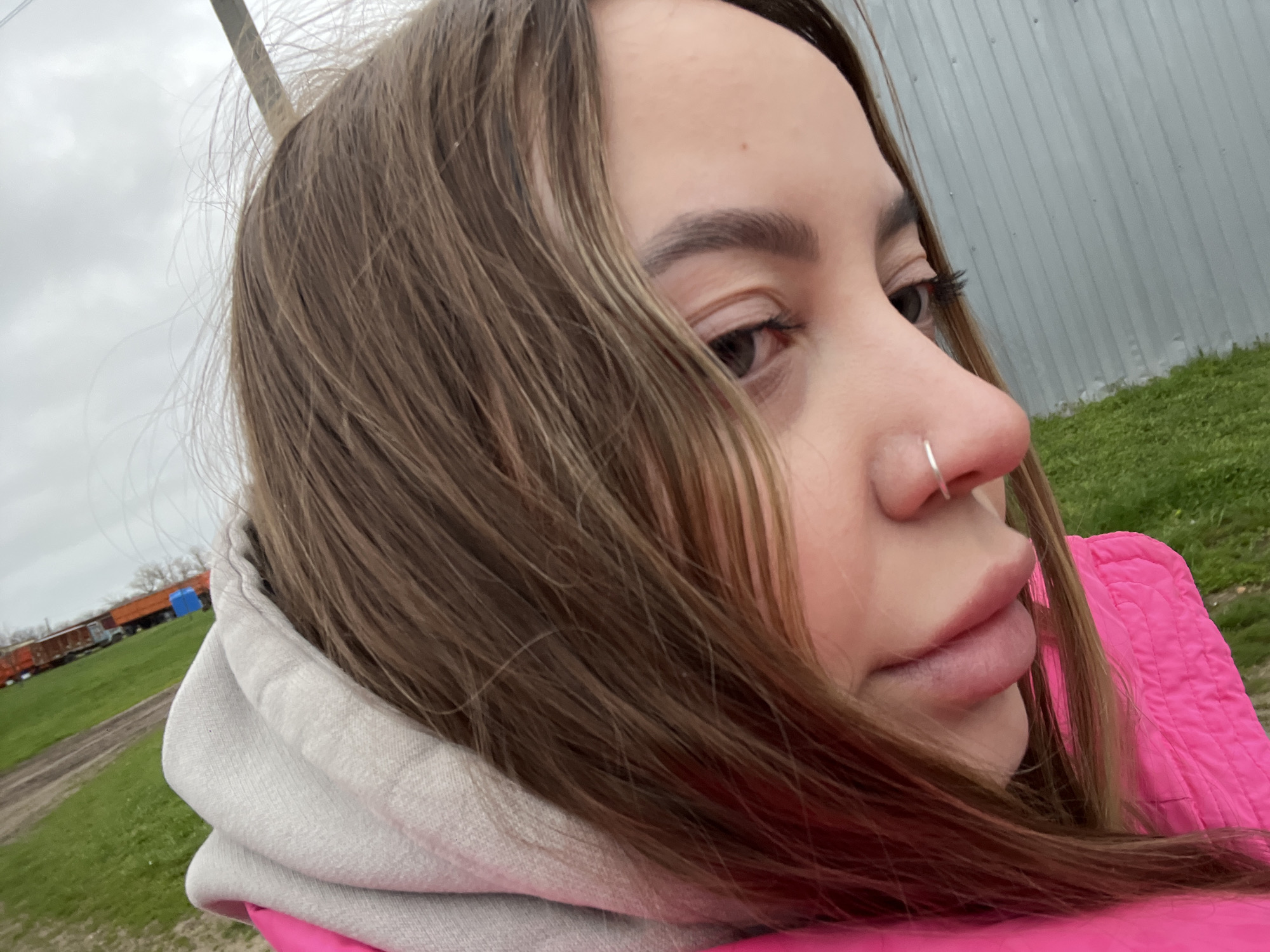 A close-up photograph shows a young woman with brown hair and a silver nose ring. She is wearing a gray hoodie underneath a bright pink jacket. The background features a gray metal fence, green grass, and a distant freight train. The photo was likely taken in an outdoor setting, possibly on a cloudy day in Novopokrovskaya, Russia.
