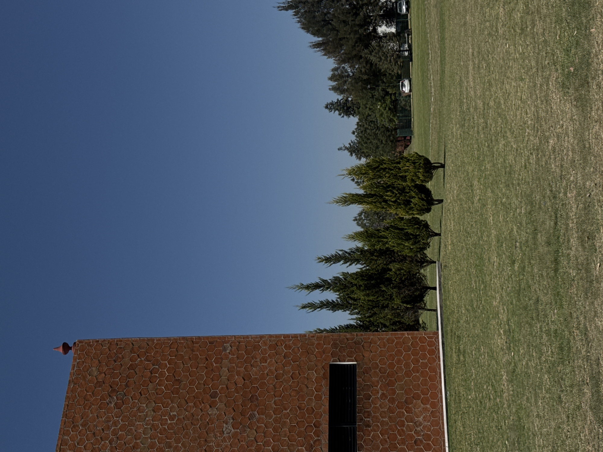 This image captures an expansive outdoor scene in San Andrés Cholula, Mexico, under a clear, bright blue sky, indicating sunny weather. The photograph is oriented vertically, but when viewed as if rotated 90 degrees counter-clockwise, it shows a wide landscape.
On the left side, a building with a distinctive facade made of reddish-brown, hexagonal-patterned tiles or bricks is visible. A small, pointed, reddish finial or spire tops the building's peak on the left. Towards the lower center of the building's wall, there's a dark, rectangular opening, possibly a window or vent, featuring vertical grilles.
Running across the middle ground, separating the sky and the building from a large field, is a line of evergreen trees. These trees are relatively tall and dense, providing a natural barrier. Beyond them, a vast, neatly manicured grassy field extends across the right side of the frame, suggesting a park, golf course, or large lawn. In the distant background, behind the field and more dense tree lines, several vehicles (appearing to be cars or golf carts) are partially visible, hinting at a nearby road or parking area.
The scene is tranquil and devoid of human activity, conveying a sense of peaceful openness. The time of day appears to be mid-morning or early afternoon, given the clear sky and the angle of the sunlight. No visible text is present in the image.