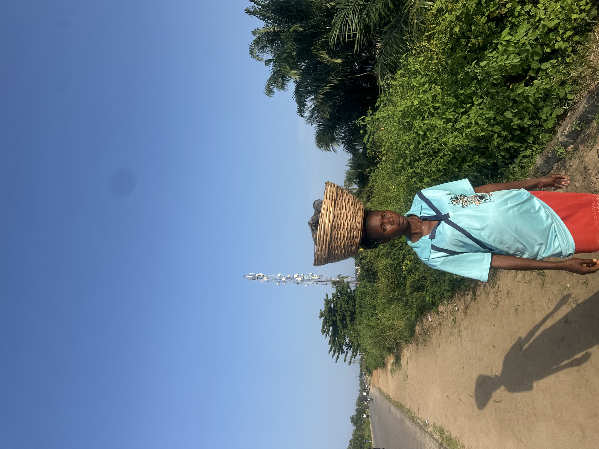 In Ganyingbo, Nigeria, a woman walks along a dirt path next to a paved road under a clear blue sky. She carries a large woven basket filled with dark objects on her head. She wears a light blue polo shirt with a dark blue stripe across the chest and a red skirt. The path is lined with lush green vegetation, and palm trees are visible in the background. A tall telecommunications tower stands in the distance. Several motorcycles are on the road, and a person on one of them is visible in the distance. The woman's shadow is cast on the path to her left.