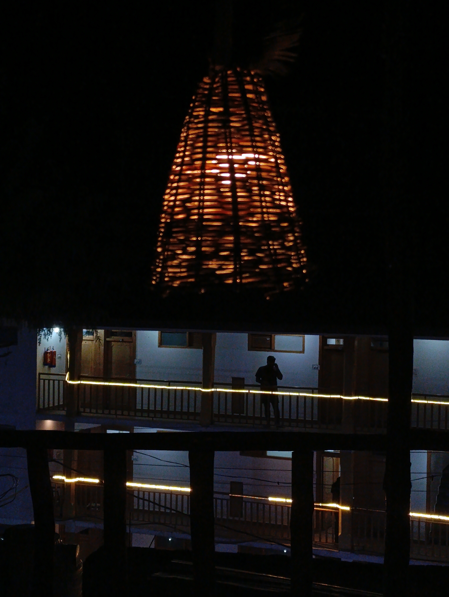 This media file depicts a dimly lit nighttime scene, likely in Vashisht, India. The primary subject is a conical, woven lampshade hanging from the ceiling, emitting a warm, orange glow from an internal light source. Below the lampshade, a multi-story building is visible, featuring balconies and doorways.
One person, silhouetted against the light of the building, is standing on an upper balcony, seemingly looking at their phone or in conversation. This person is the only clearly discernible human figure. The building's architecture suggests an outdoor courtyard or passage, with wooden railings and posts forming the balconies. Yellow string lights are visible along the railings of the balconies, adding to the ambient illumination.
The overall impression is one of a tranquil, possibly tourist-oriented lodging or guesthouse at night. The dark background and the focused light from the lampshade and string lights create a sense of depth and intimacy. There is no visible text in the image. The time of day is clearly night, and the weather appears to be clear. The scene evokes a feeling of calm and relaxation.