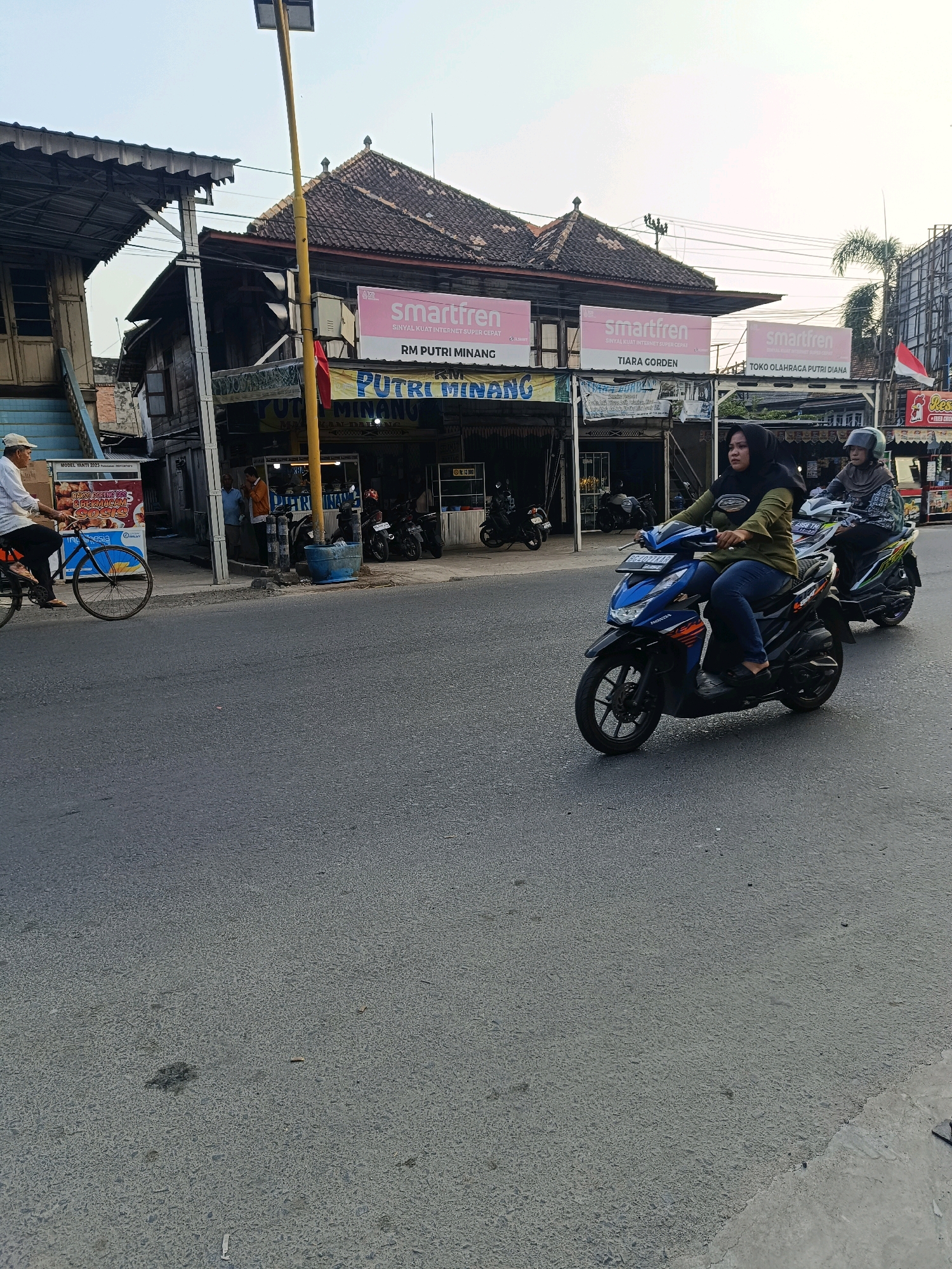 The image depicts a street scene in Kayu Agung, Indonesia, at approximately 5:30 PM. The time is estimated based on the warm, soft lighting and the shadows cast. The scene captures a mix of commercial buildings and street activity.
In the foreground, a paved road occupies the majority of the frame. The road is asphalt and appears well-traveled. Two motorcycles are moving along the road. The lead motorcycle is blue and is being ridden by a woman wearing a green hijab and blue jeans. She is looking forward. Behind her, another motorcycle is partially visible, ridden by a person wearing a light-colored hijab and a patterned top, also looking forward.
On the left side of the road, a man wearing a white shirt and a cap is riding a bicycle. He is heading towards the left of the frame. The bicycle is a dark color.
The background features several buildings. The central building is a two-story structure with a dark-tiled roof. It has multiple signs advertising "smartfren" and other businesses, including "RM PUTRI MINANG" and "TIARA GORDEN." One sign also mentions "TOKO OLAHRAGA PUTRI DIANA." The building's facade shows signs of age, with weathered wood and some exposed structural elements. Below the upper story, there are storefronts, some with open entrances, suggesting commercial activity. Several motorcycles are parked in front of these establishments.
To the left of the central building, another structure with a corrugated metal roof is visible, appearing to be an extension or a separate shop. A red flag is hanging from a pole near the entrance.
To the right of the central building, another building is partially visible with a red and white Indonesian flag attached to its side. Signs for "Resto" and other food-related businesses are present. Further down the street, other buildings and what appear to be utility poles are visible. The sky is a clear, pale blue, indicative of late afternoon. The overall impression is a busy, typical street in a Southeast Asian town.