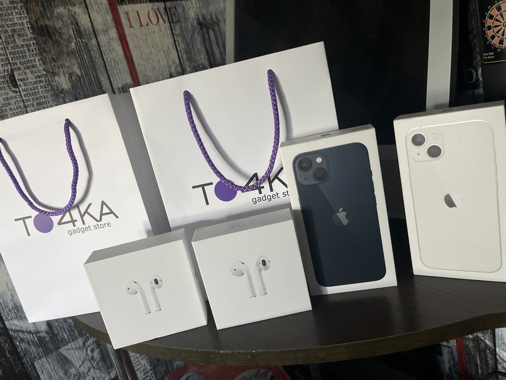 This image displays a collection of Apple products and branded shopping bags arranged on a dark wooden surface. The primary subjects are two white boxes, each depicting a pair of white wireless earbuds, likely AirPods, and two iPhone boxes. One iPhone box is dark blue and shows a dark blue iPhone inside, while the other is white and displays a white iPhone. The background features two white shopping bags with purple handles, each bearing the "TO4KA gadget store" logo. Faintly visible in the upper left and right of the image are parts of a textured wallpaper with text and a dartboard, respectively. The scene appears to be indoors, likely in a retail or personal collection setting. The lighting suggests a well-lit environment, possibly during daytime, though no specific time or weather conditions are indicated. The overall presentation is neat and organized, highlighting the products. The visible text includes the store name "TO4KA gadget store," "I LOVE," and some fragmented text on the wallpaper and dartboard. The location context of Bohunove, Ukraine, is provided, but there are no explicit visual cues within the image to confirm this specific location.