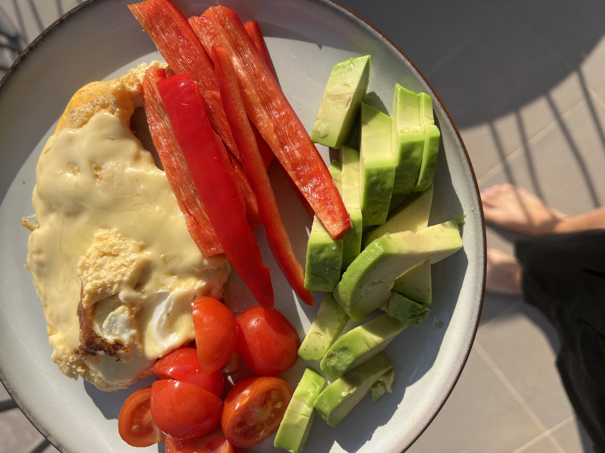 The image displays a close-up, overhead view of a healthy meal served on a light-colored plate, set outdoors on what appears to be a sunny morning in Budva, Montenegro.
The main subject is a plate of food. On the left side, there's a portion of fluffy, light yellow eggs, possibly scrambled or a folded omelet, generously topped with a layer of smooth, melted, creamy yellow cheese. Below this, at the bottom left of the plate, are several bright red cherry tomatoes, cut in halves.
Filling the middle and right sections of the plate are fresh vegetables. A pile of vibrant red bell pepper strips, cut lengthwise, is arranged next to the eggs and cheese. To the right of the bell pepper, numerous chunks of fresh, green avocado are neatly placed. The food items appear fresh and colorful.
The scene is set outdoors, likely on a balcony or patio, indicated by the bright, natural sunlight casting strong, distinct shadows both on the plate and on the light-colored tiled floor in the background. The shadows of a railing or window frame are visible on the floor. In the blurred background, parts of a person's bare feet and lower legs are visible on the tiled surface, accompanied by a dark garment, suggesting someone is seated nearby enjoying the meal. The clear, bright light suggests a morning or early afternoon setting. No text is visible in the image.
