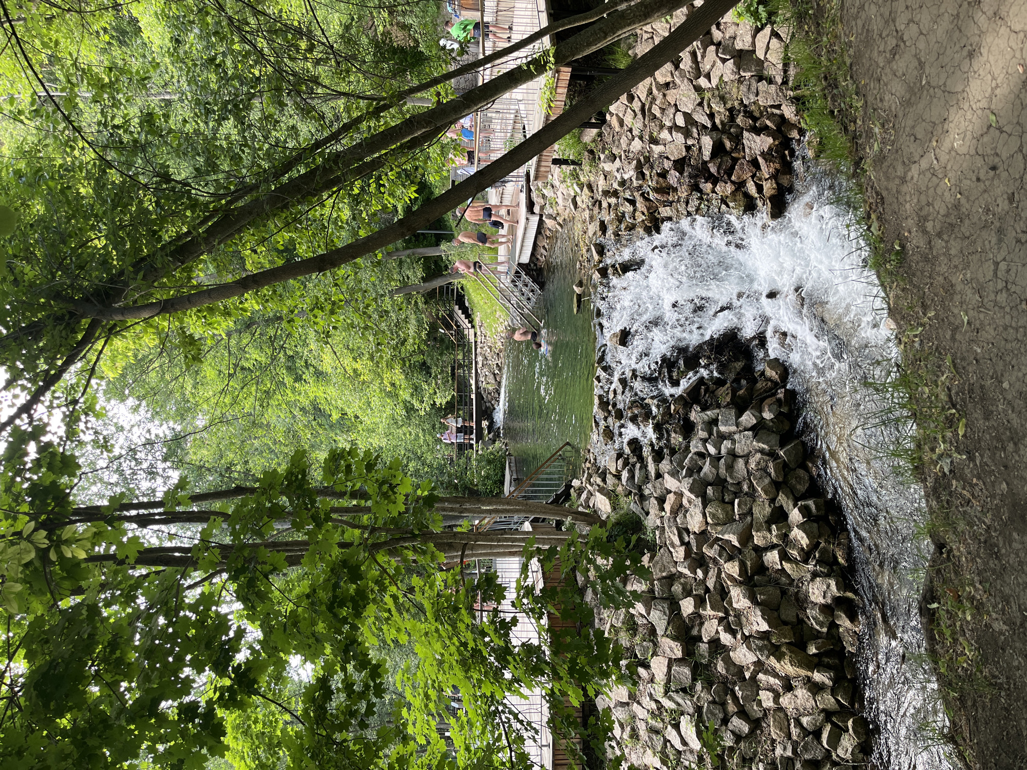 The image depicts a scenic outdoor area, likely a park or natural reserve, with a focus on a water feature. In the foreground, a small waterfall cascades over a rocky embankment into a stream. The water is clear and appears to be flowing rapidly. Surrounding the water are natural elements, including large, rough-hewn rocks and lush green vegetation. Wooden walkways and platforms are visible along the banks of the stream and a larger body of water, suggesting this is a developed recreational area. Several people are present, some of whom are in the water, indicating swimming or water-based activities. Others are standing on the walkways, observing the scenery or interacting with each other. The overall environment is verdant, with numerous trees providing a canopy of green leaves. The lighting suggests it is daytime, with sunlight filtering through the trees.
This location is likely the Gorky Park in Kazan, Russia. The park is known for its natural beauty, including its riverfront areas and walking paths, and often features recreational activities like swimming in designated areas during warmer months. The presence of walkways, clear water, and people engaged in leisure activities are consistent with the typical appearance of the park.