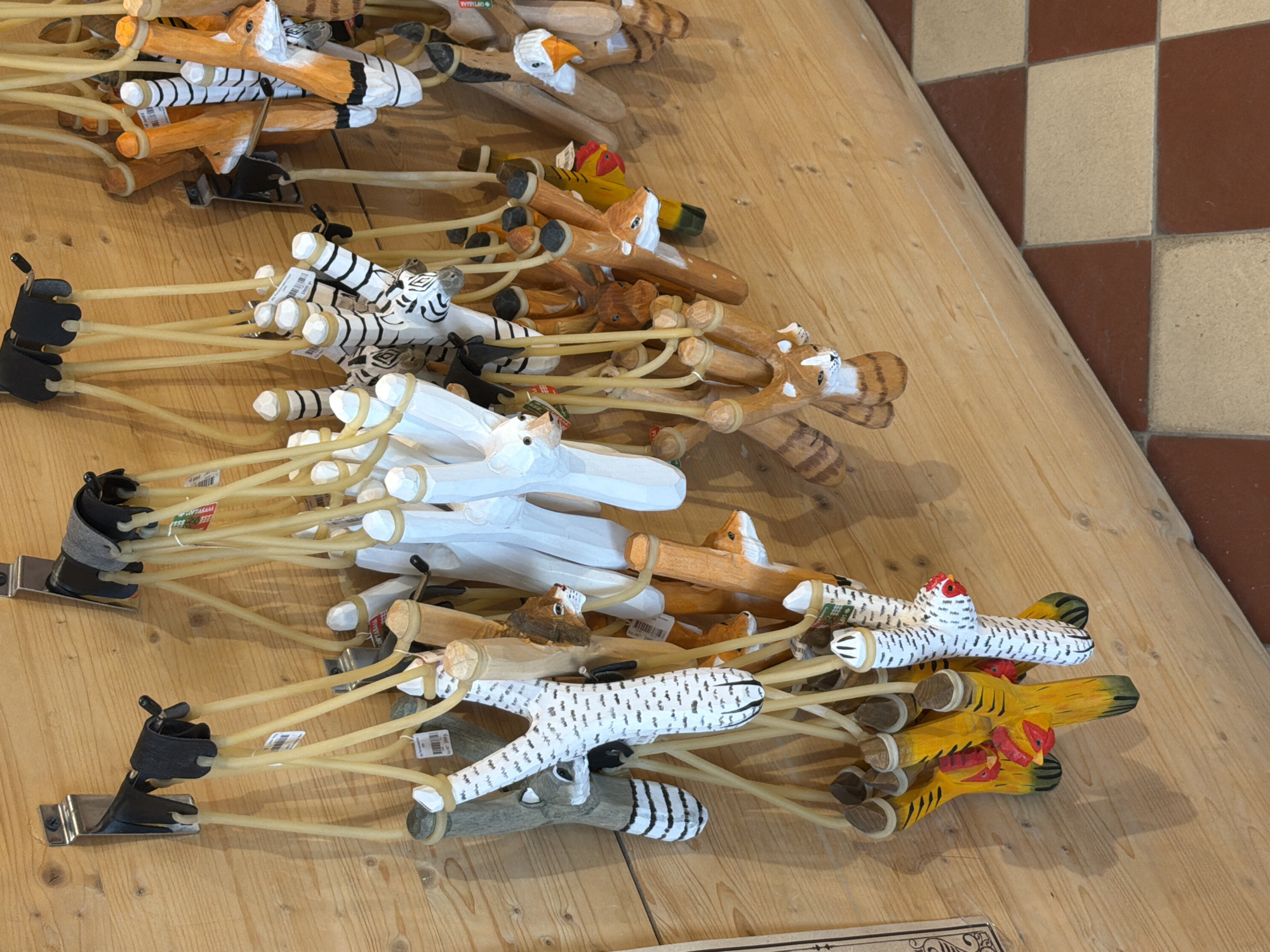 The image shows a display of handcrafted wooden slingshots, each carved and painted to resemble various animals. These are hanging from metal hooks mounted on a light-colored wooden wall. The slingshots have yellow elastic bands. Among the animals represented are a white polar bear, a striped zebra, a fox, a deer, a chicken, a racoon, and a cat. Some slingshots have small price tags attached. The flooring visible at the bottom of the image is a checkerboard pattern of brown and beige tiles. This scene is consistent with a small shop or market stall, potentially in Sortavala, Russia, a city known for its arts and crafts.