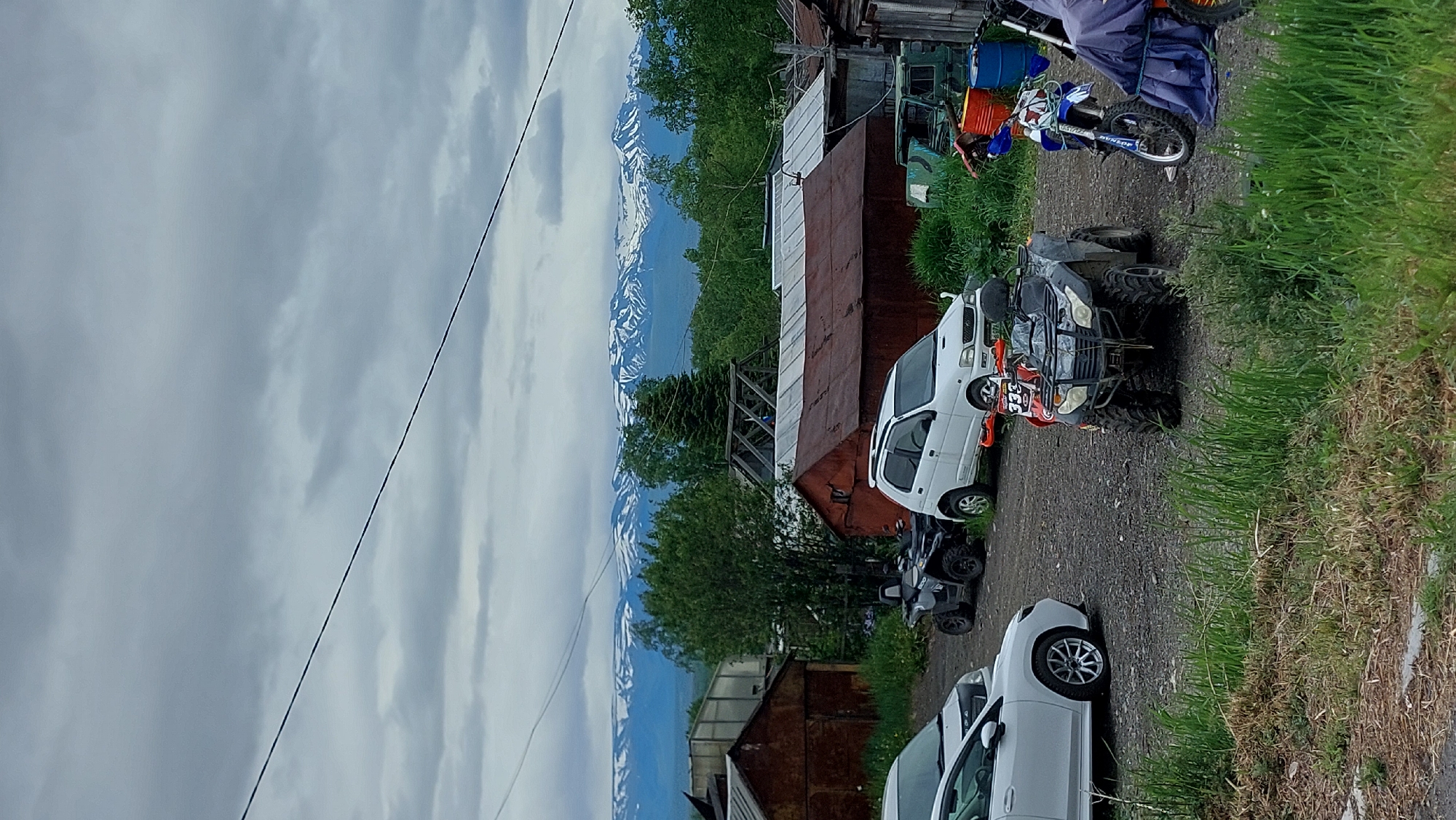 The image depicts a scene in an outdoor, rural area, possibly the city of Zelenyy, Russia, with a backdrop of snow-capped mountains. In the foreground, a gravel lot is occupied by several vehicles including a white sedan, a black ATV, a white compact SUV, a red ATV, and a dirt bike. There are also several dilapidated wooden structures, likely sheds or outbuildings, with rusty metal roofs. The vegetation consists of tall grass and some trees. The sky is overcast with gray clouds. The overall impression is one of a remote, perhaps slightly unkempt, residential or workshop area.