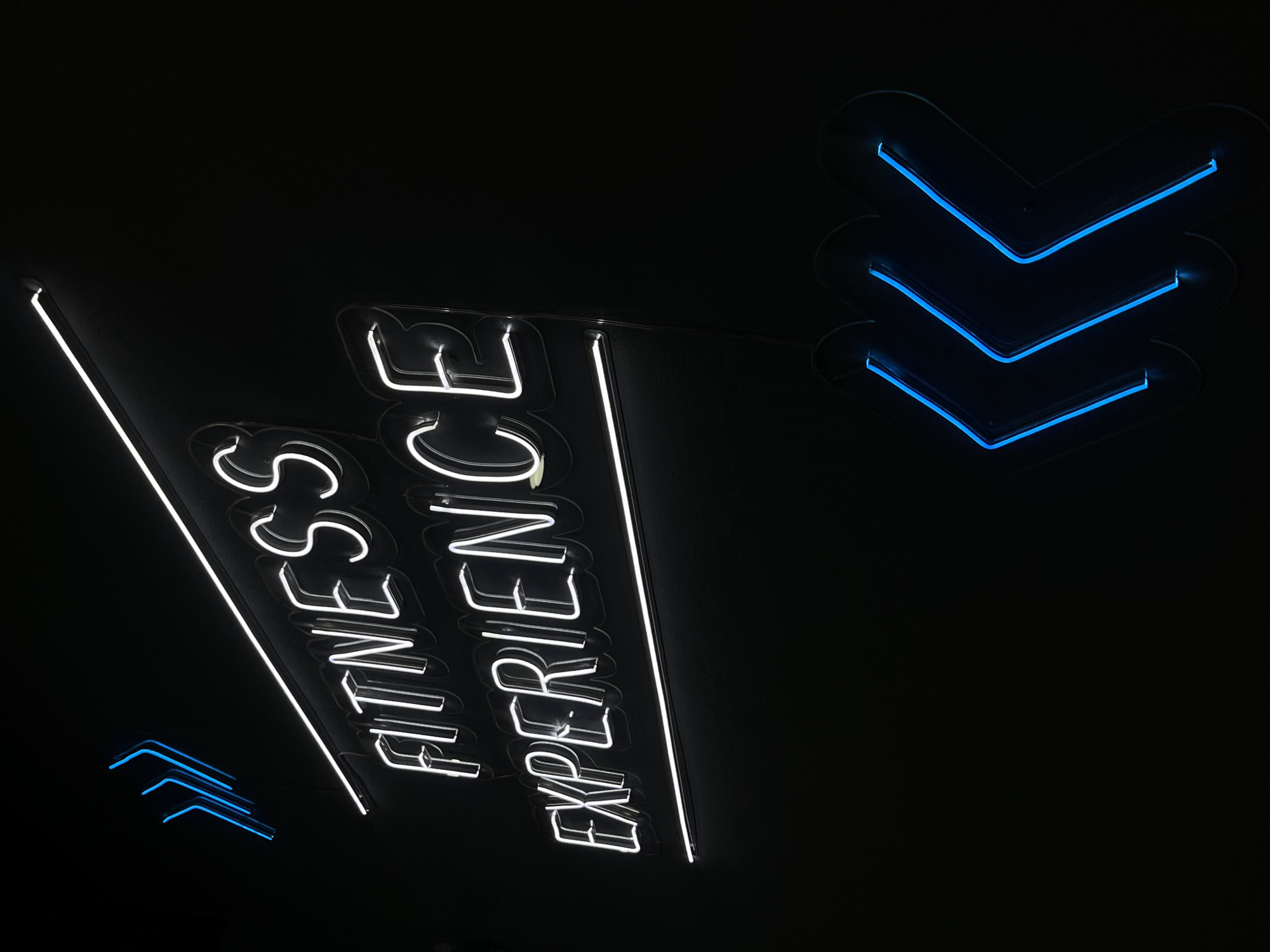 The image shows a sign in a dark setting. The sign reads "Fitness Experience" in white neon lettering. To the right and left of the text are clusters of blue neon arrows pointing to the right. The location is Unknown, Unknown.