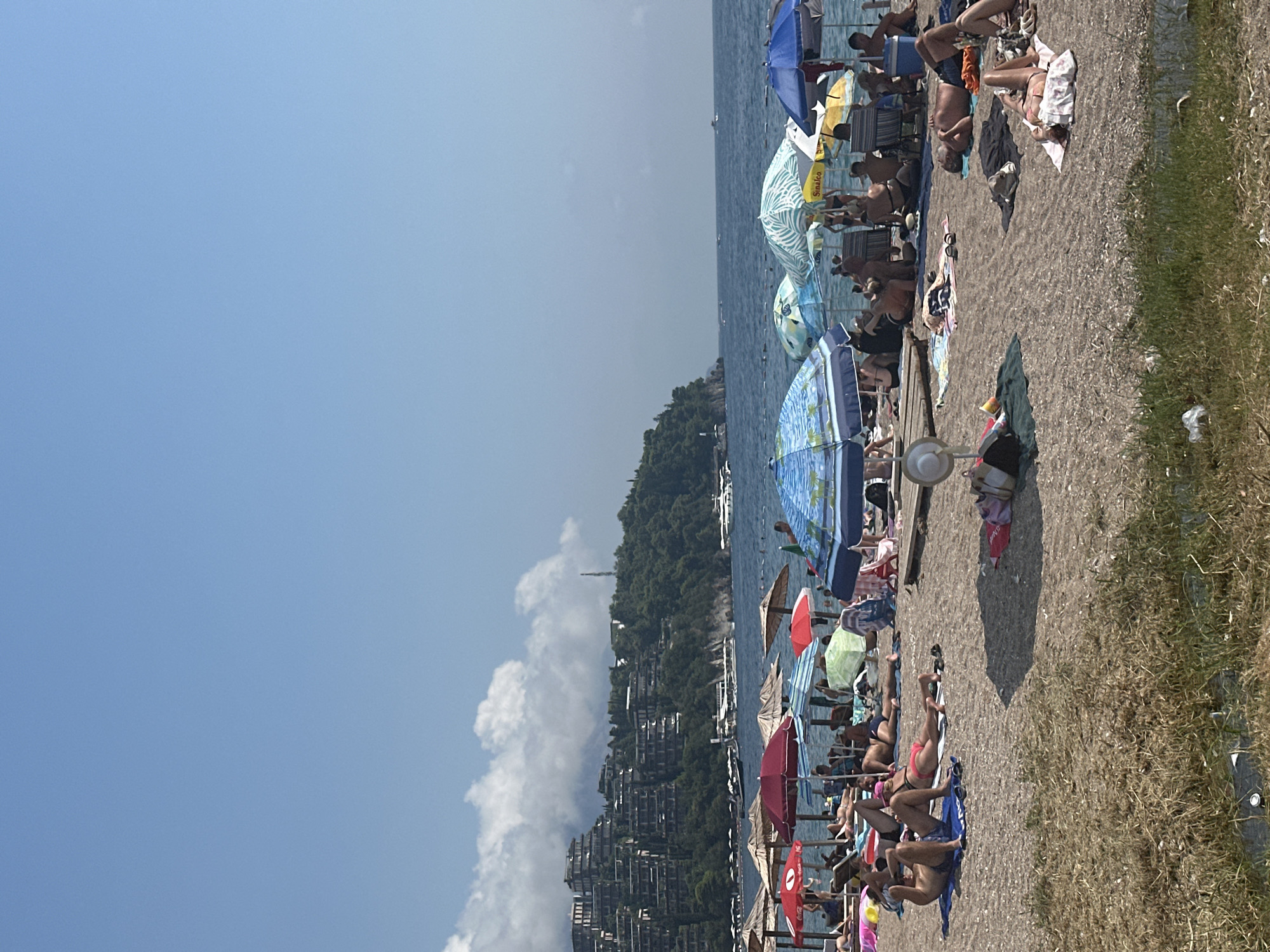 The image depicts a crowded beach in Budva, Montenegro, on a sunny day. Numerous people are sunbathing on towels and chairs scattered across the pebbly shore. A variety of colorful umbrellas, some patterned and some solid, provide shade. In the background, the turquoise Adriatic Sea stretches out, with a few boats visible in the distance. Behind the beach, a lush green hillside is dotted with tiered buildings, characteristic of Budva's coastal architecture. The sky above is a clear, pale blue, with a few scattered white clouds towards the horizon.