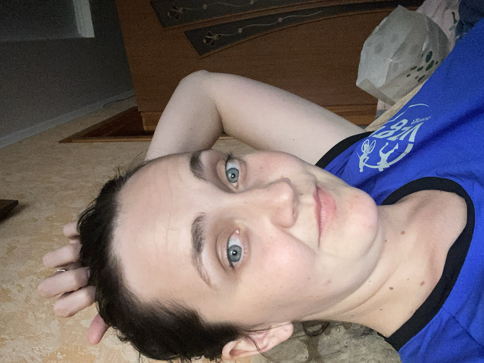 In this close-up, indoor, selfie-style photograph, a woman with light skin, blue eyes, and dark hair pulled back is the central subject. She is wearing a blue t-shirt with a black collar. Her left arm is raised and bent, with her hand resting on the back of her head, while her right hand is on top of her head. Her expression is neutral, with a slight smile. The background consists of a textured wall and a wooden cabinet. The lighting is dim, suggesting an indoor environment, likely a bedroom or living area. The photograph is taken at a slight upward angle, focusing on the woman's face and upper body. There is no visible indication of a city or specific location.