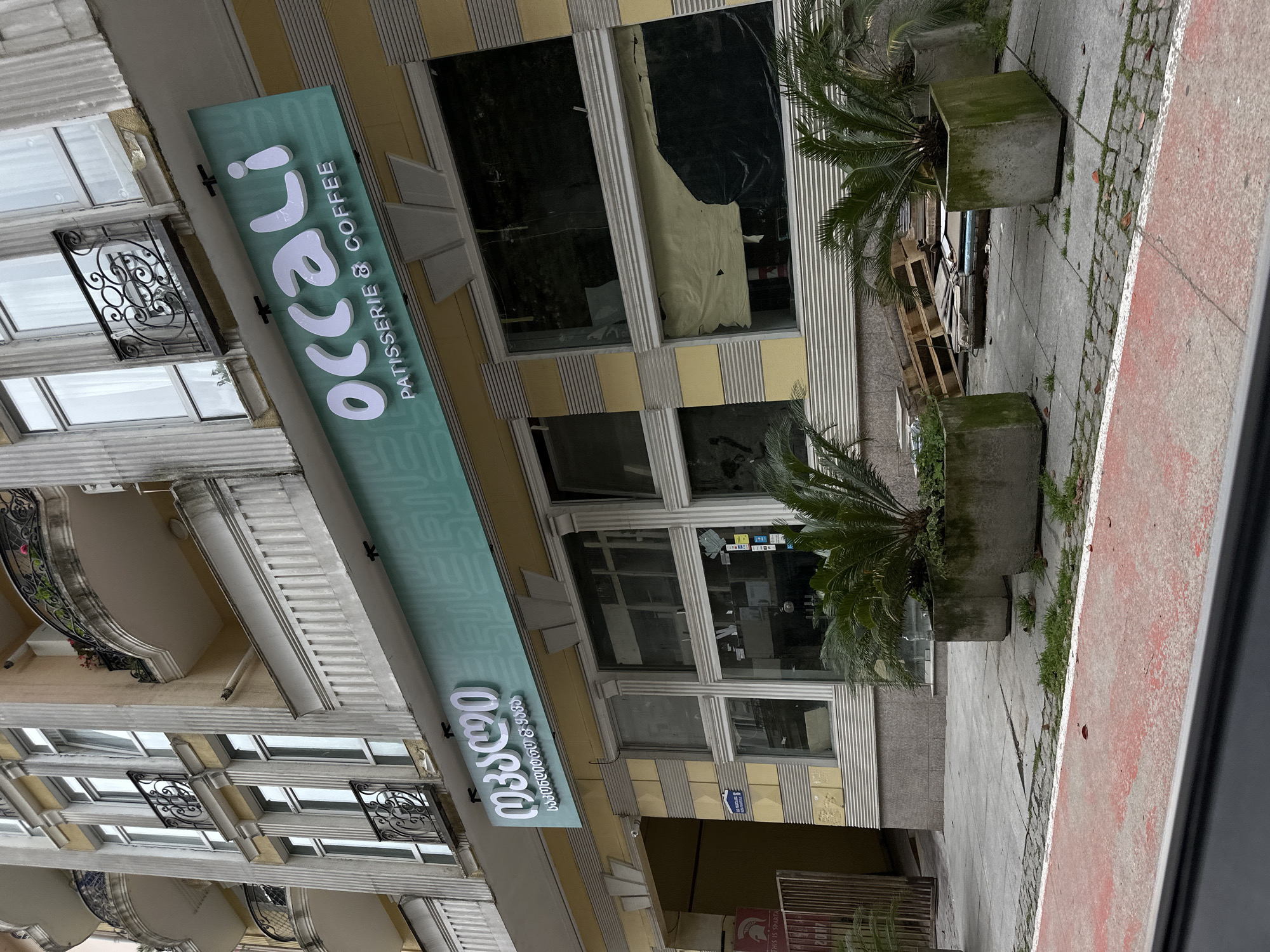 The image depicts the exterior of a building, likely a commercial establishment, situated in Batumi, Georgia. A sign for "Occeli Patisserie & Coffee" is prominently displayed in both Georgian and English, indicating the type of business. The building features a multi-story facade with windows, balconies, and decorative architectural elements. Two potted palm trees are placed on the sidewalk in front of the building. The sidewalk appears to be made of concrete and brick, with a red painted bike line visible. A stack of wooden pallets is placed near the building.