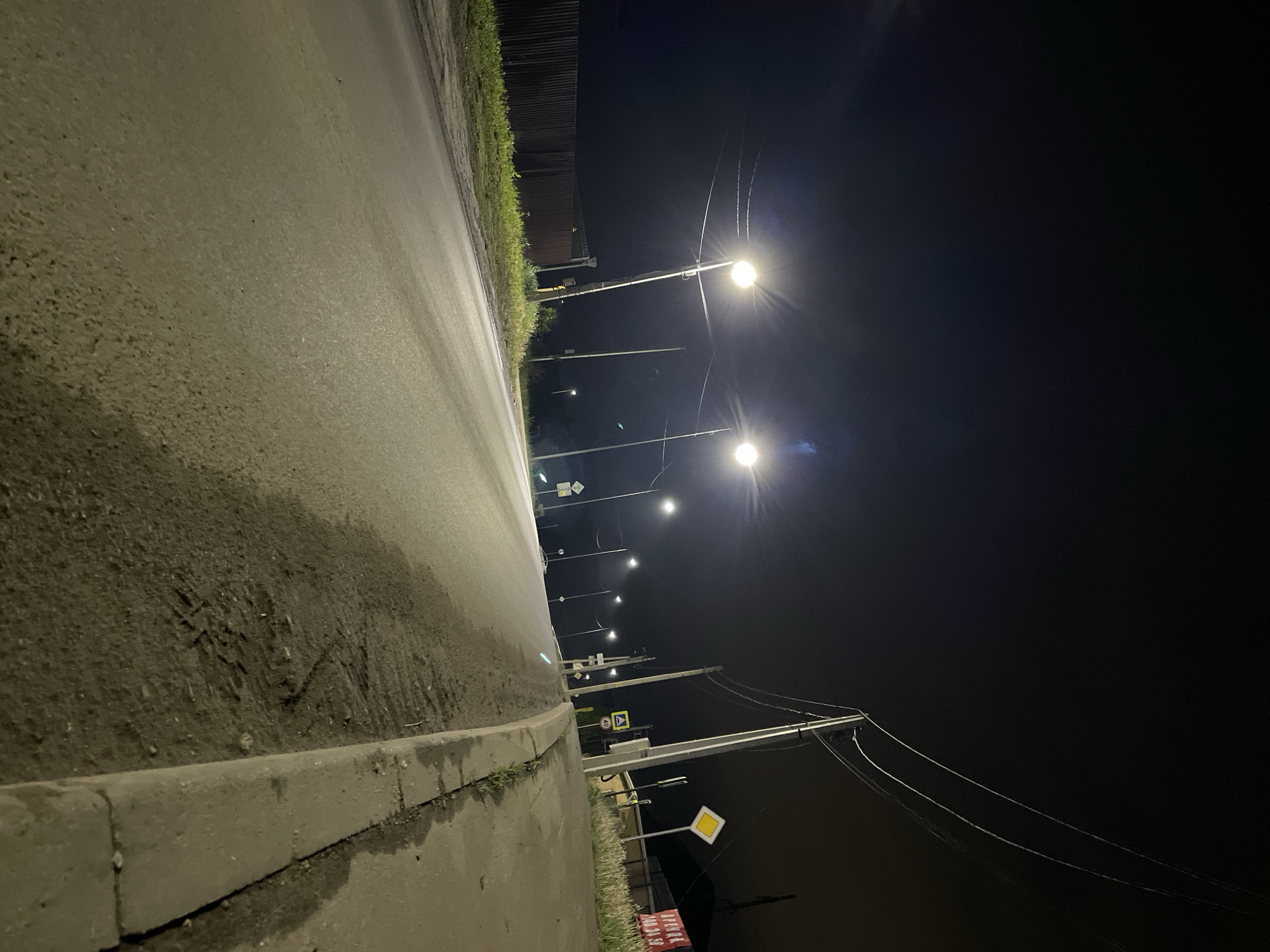A night-time perspective looking down a street in Эстачи, Russia. The foreground is a gray asphalt street with a concrete curb on the left. Several street lights illuminate the scene, spaced along the street. The right side of the frame is dominated by darkness, with utility poles and wires visible against the night sky. Street signs can be seen along the road. Vegetation lines the edges of the road.
