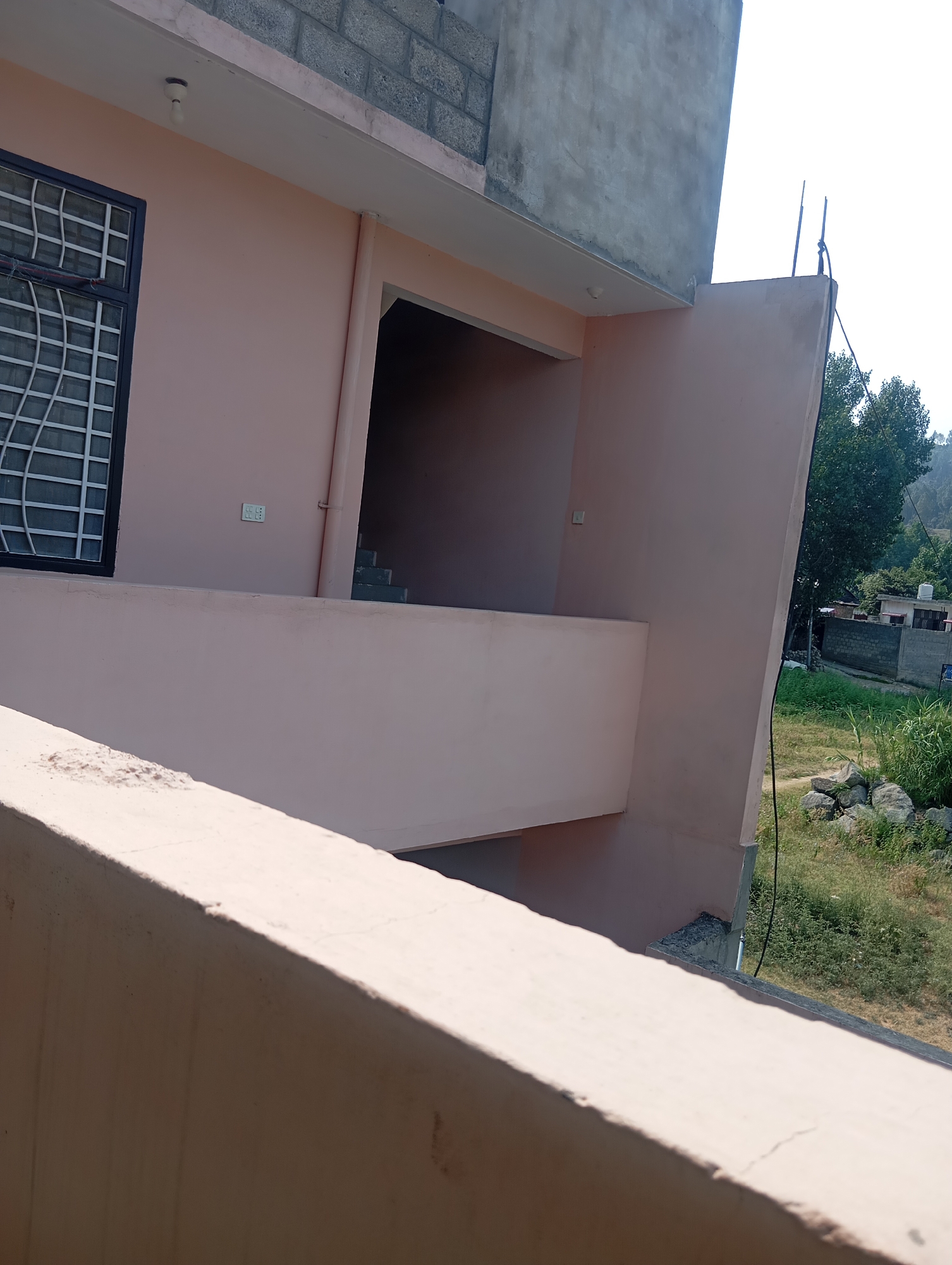 This is a medium-angle outdoor shot of a building under construction or renovation, likely in a rural or mountainous area, given the greenery and distant hills visible in the background. The building's facade is primarily painted a light pink, with sections of rough, gray concrete. A dark-framed window with a white metal grate is visible on the left side of the image.
A balcony or ledge, also painted pink, extends across the lower portion of the building. Part of a similar structure is in the foreground, suggesting the photo was taken from another balcony or elevated viewpoint. Above the pink facade, there's a section of exposed gray concrete wall and a tiled roof.
A dark opening reveals stairs leading upwards inside the building. An electrical outlet with two sockets is visible on the pink wall near the stairs. A simple light fixture with a bare bulb hangs from the ceiling near the top left of the image.
The sky is bright and clear, indicating it is daytime, likely with fair weather. The presence of scaffolding-like structures and unfinished concrete suggests ongoing construction work. The overall scene conveys a sense of a work in progress in a natural, possibly developing, environment. There is no visible text in the image.