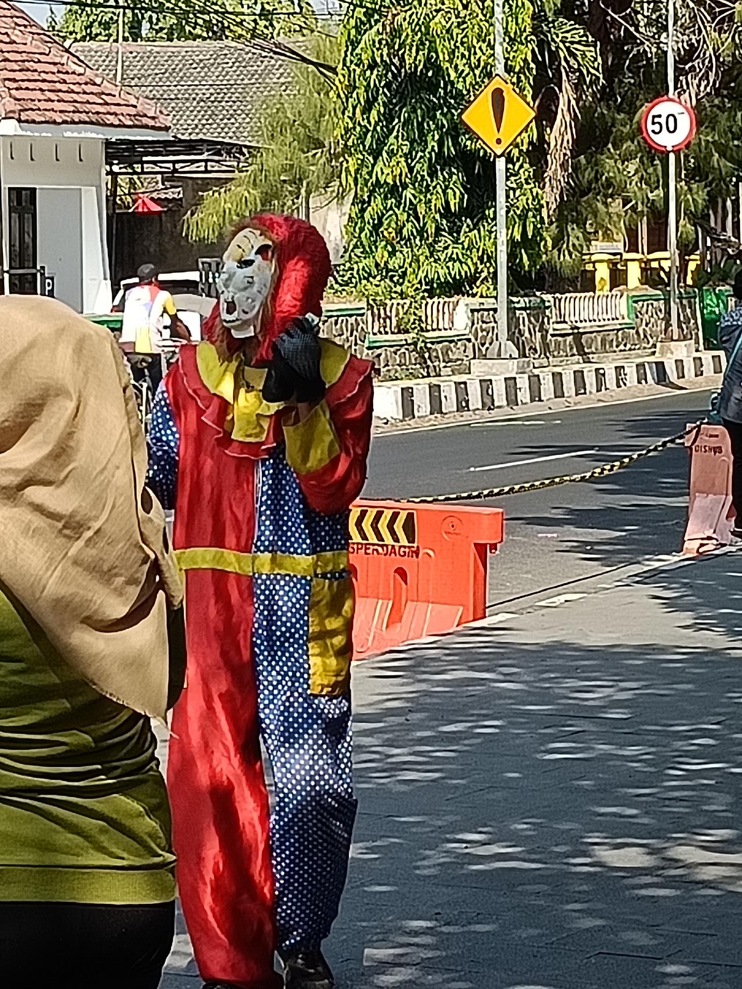 A person in a clown costume with a red and yellow ruff, a white and red mask, and a red and blue polka dot suit stands on a street in Kediri City, Indonesia. The clown is wearing black gloves and black shoes. In the background, there is a yellow diamond-shaped sign with a black exclamation point, and a circular speed limit sign indicating 50. Orange traffic barriers are placed on the side of the road, and a yellow and black striped caution tape is stretched between them. The street is paved, and there are shadows cast by trees on the surface. A portion of a building with a tiled roof is visible to the left. Another person wearing a beige headscarf and a green top is partially visible in the foreground.