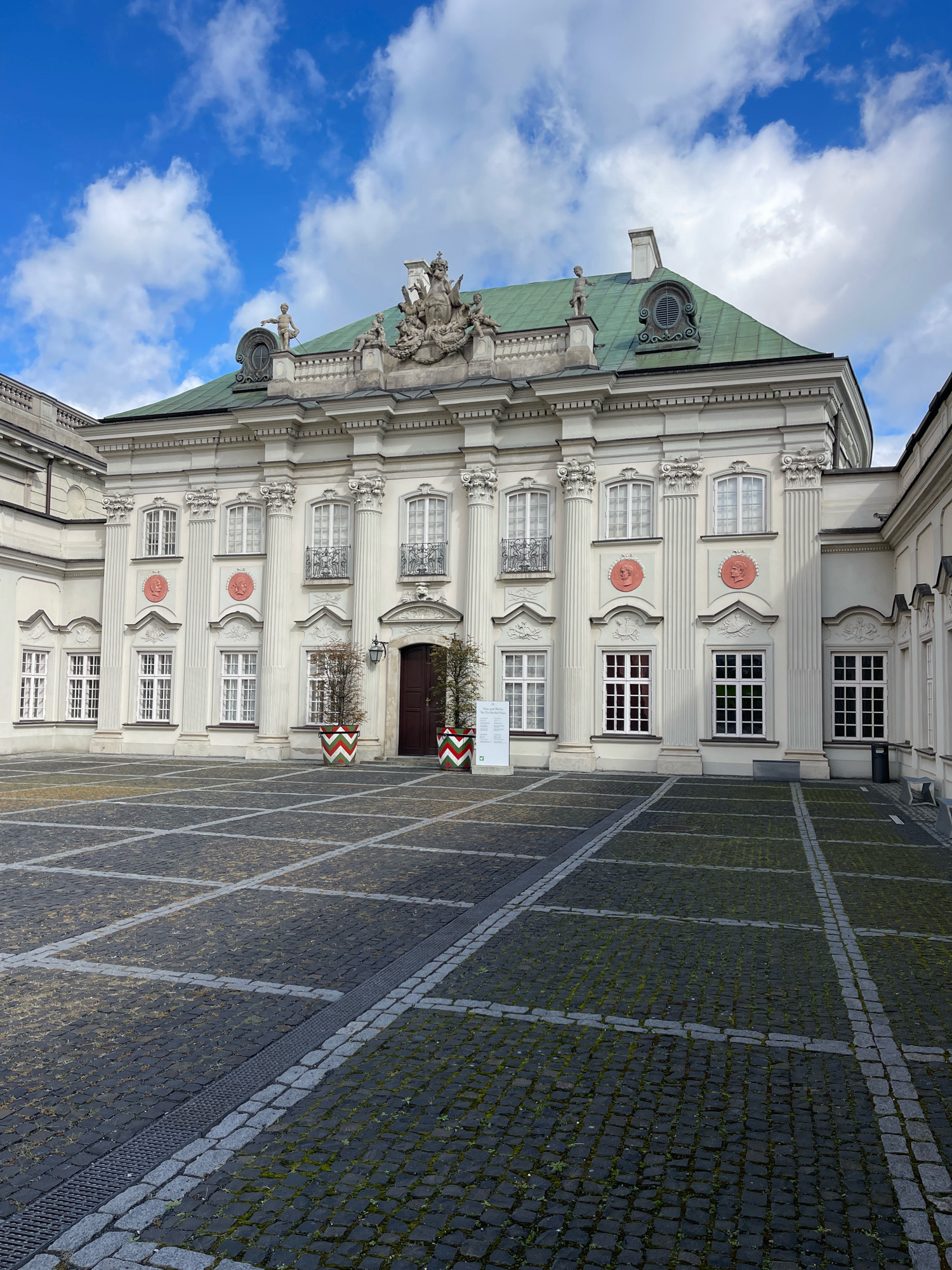 The image shows the exterior of the Wilanów Palace in Warsaw, Poland. The palace is a Baroque-style residence, notable for its white facade, green roof, and ornate architectural details. In the foreground, a cobblestone courtyard is visible, with a distinct grid pattern formed by lighter colored stones. Two large planters with a red and green chevron pattern are placed near the entrance of the palace. The sky above is blue with scattered white clouds. No people are visible in the image. The overall scene conveys a sense of grandeur and historical significance.