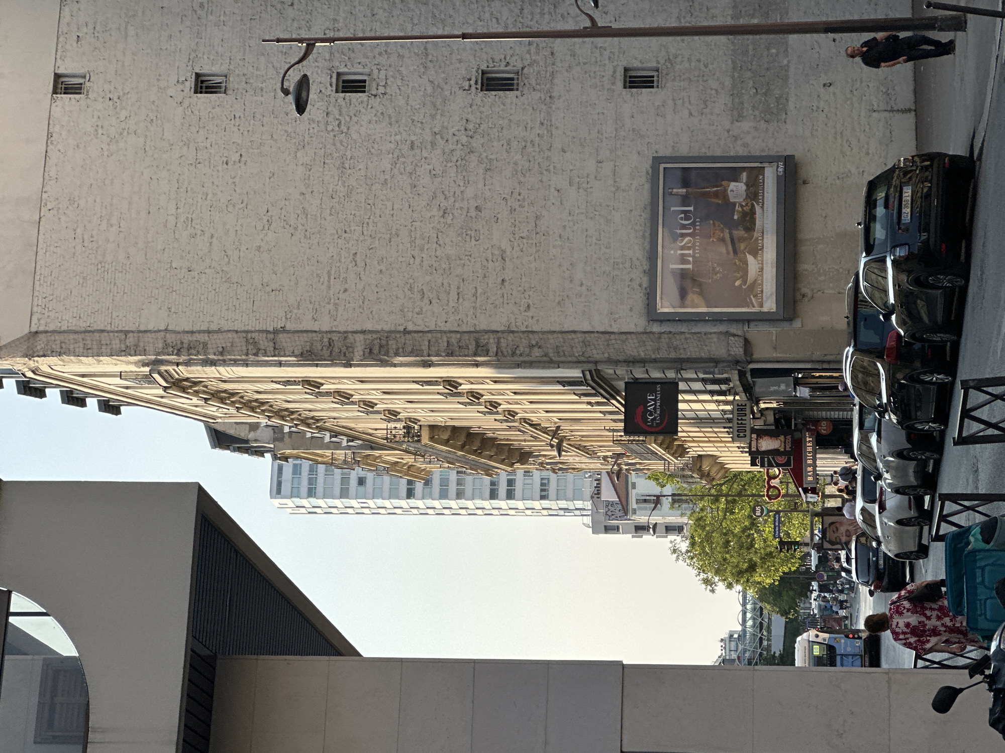 This is a street-level, medium shot of a street in Paris, France, captured in daylight. The perspective is looking down the street, with buildings lining both sides. The left side of the frame is dominated by a modern, light-colored building with a curved overhang and dark horizontal blinds visible beneath it. On the right side, an older, multi-story building with a textured, light-colored brick facade is prominent. Several windows with barred openings are visible on this building.
A large advertisement for "Listel" is displayed on the right-hand building, featuring a bottle and glasses. A lamppost with a distinctive curved arm stands near the advertisement. The street itself is paved and appears to be a one-way street. Several cars are parked along the right curb, including a black SUV with license plate GZ 358 LT. Pedestrians are present on the sidewalk, with one man in a dark shirt and jeans walking away from the camera on the right.
Further down the street, more buildings with balconies and ornate architectural details are visible. Signs for "LA CAVE ENTREPRENEURS" and "COIFFURE" are mounted on the buildings. Traffic, including buses and other vehicles, is present in the distance. A woman in a floral print shirt is walking away from the camera in the foreground, with a blue bin bag beside her. A motorcycle is parked in the lower left corner. The sky is clear and light. The overall impression is a bustling urban street scene.
