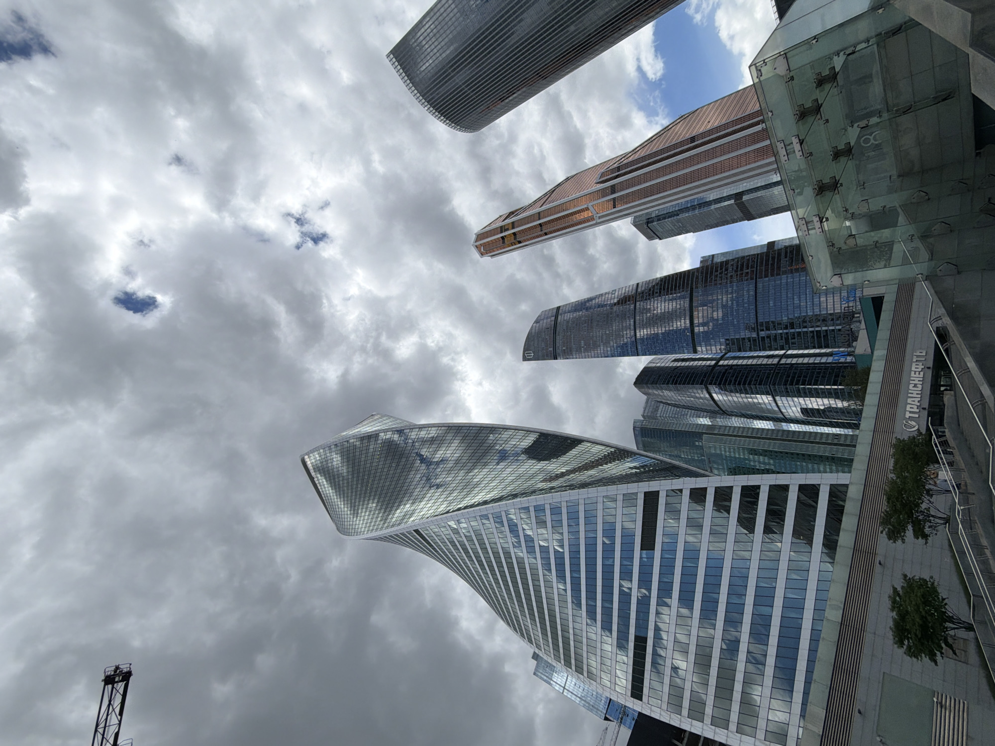 The image shows a low-angle view of several skyscrapers in the Moscow International Business Center (also known as Moscow City) in Moscow, Russia. The sky is filled with clouds. The most prominent building is the Evolution Tower, characterized by its twisting shape. Other skyscrapers are partially visible in the background. There are trees and the word "ТРАНСНЕФТЬ" on one of the adjacent buildings. A construction crane is visible in the bottom left.