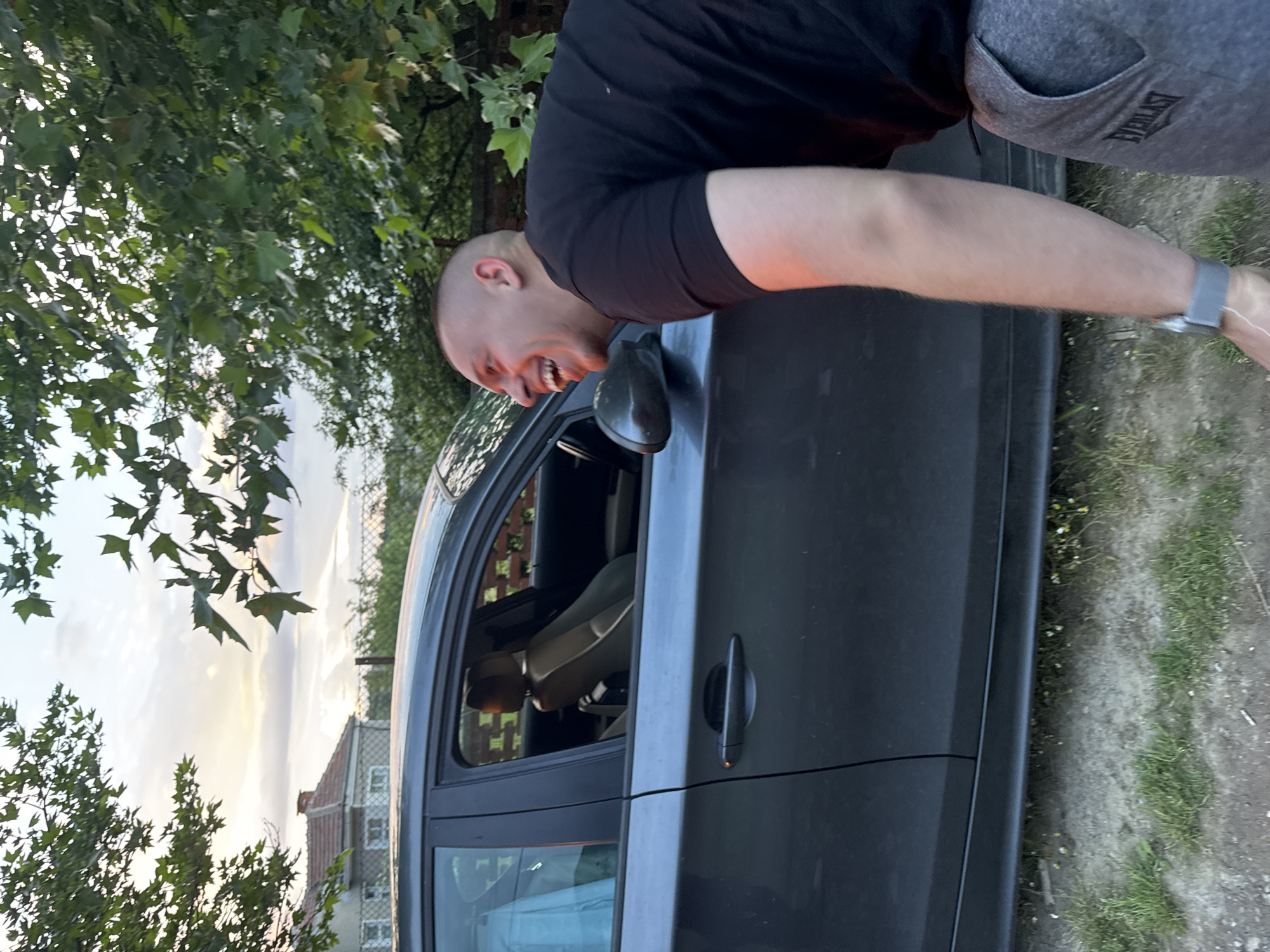 A man with a shaved head, wearing a black t-shirt and gray sweatpants, is bent over a dark-colored car. He is laughing and leaning into the open driver-side window. The car is parked on a dirt and grass surface. In the background, there are trees, houses, and a slightly overcast sky. No further information indicates a location in Ruzhitsa, Bulgaria.