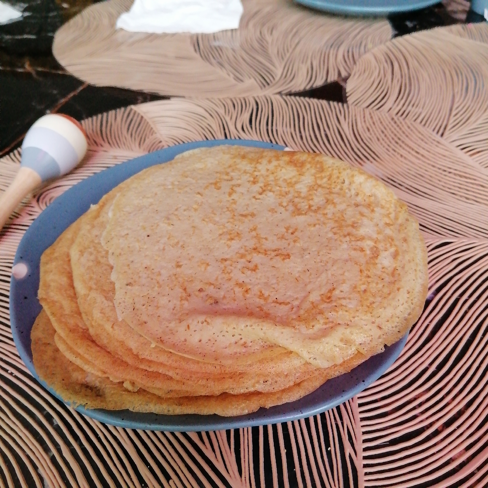 A medium-angle shot depicts a stack of pancakes on a blue plate, placed on a patterned surface. The background features a black and beige patterned cloth, with a white cloth item visible in the upper left corner. A maraca toy rests beside the plate. The image was taken in Unknown, Unknown.