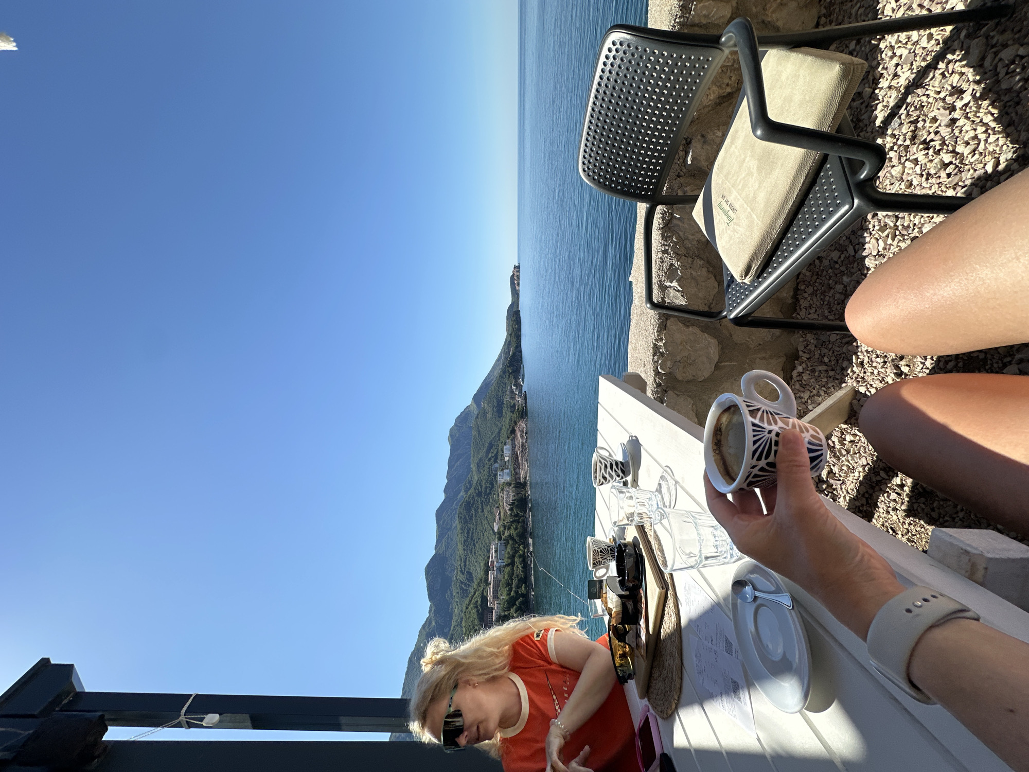 The image captures a moment at an outdoor cafe, possibly in Boreti, Montenegro, given the coastal landscape. The perspective is from someone seated at a white table laden with cups, glasses, plates, and what appears to be a menu. A hand holds a cup of coffee. The person seated across the table is a fair-haired woman wearing a red shirt and sunglasses, engaging in an action that is unclear, possibly reading the menu or using her phone.
In the background, the Adriatic Sea stretches towards the horizon, adjacent to a mountainous coastline dotted with buildings. A black metal chair with a cushion sits unoccupied near a stone wall. The setting sun casts long shadows across the scene.