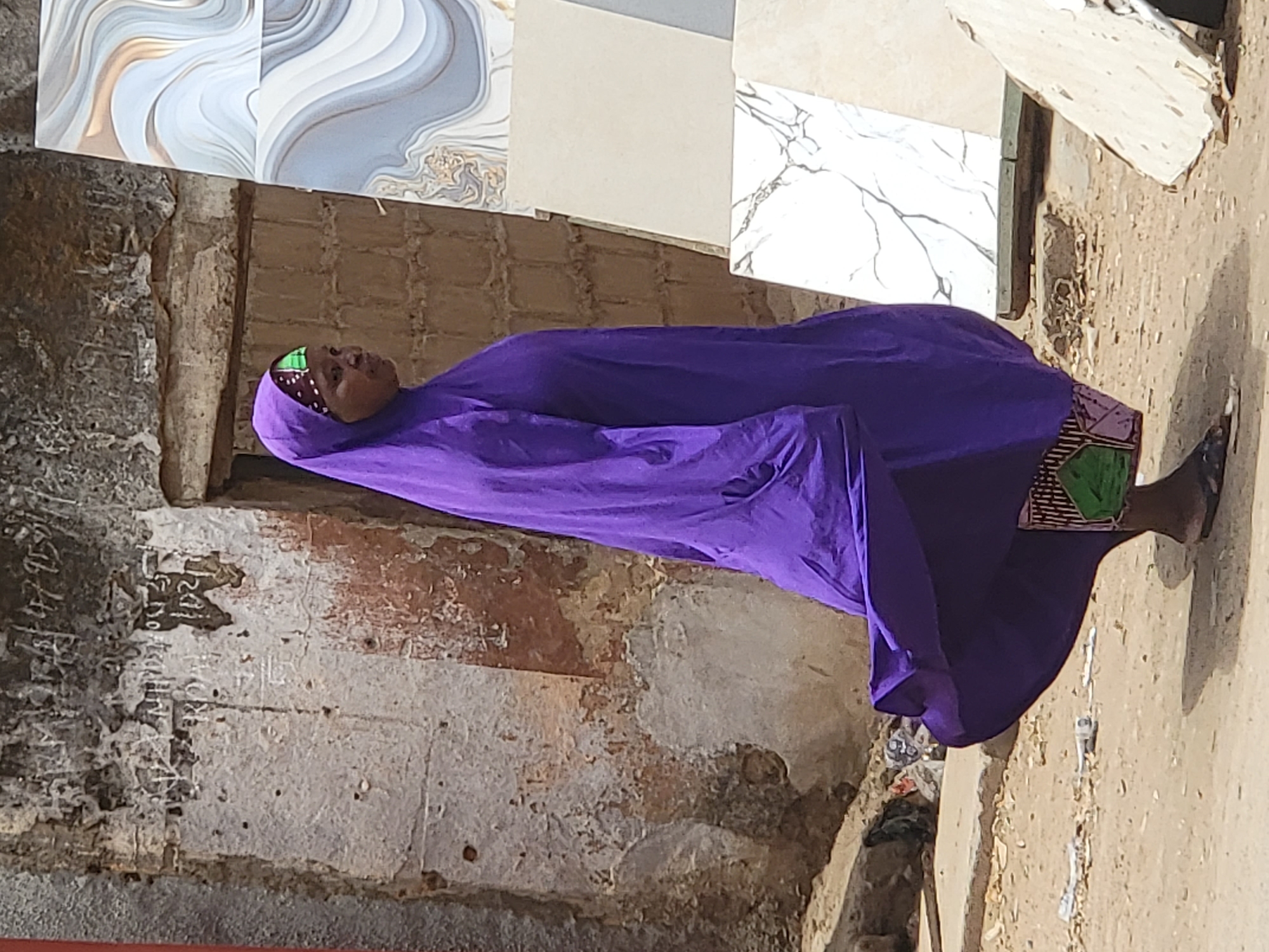 An outdoor scene in Maiburiji, Nigeria, features an individual, likely a woman, dressed in a vibrant purple full-length garment and a matching head covering, with a green and white patterned inner cap visible around her face. She has dark skin and wears dark, flat sandals. She stands looking slightly upwards and to her right, with a calm, neutral expression.
In the background, a variety of large ceramic tiles are displayed vertically, showcasing diverse designs including swirling grey and white patterns, plain light beige, and white marble with dark veining, suggesting a tile shop or construction material vendor. Below the tile display, a weathered wall made of bricks or stone is visible, with peeling plaster, faded white markings, and some illegible scribbles or partial numbers like "150" on its surface. The ground is unpaved and dusty, with some debris and a shallow ditch or gutter at the base of the wall.
The image is captured during bright daylight, indicated by strong illumination and clear shadows, implying sunny weather. The overall setting suggests a bustling, possibly older, commercial or street environment. The scene captures a candid moment of daily life without a specific event taking place.