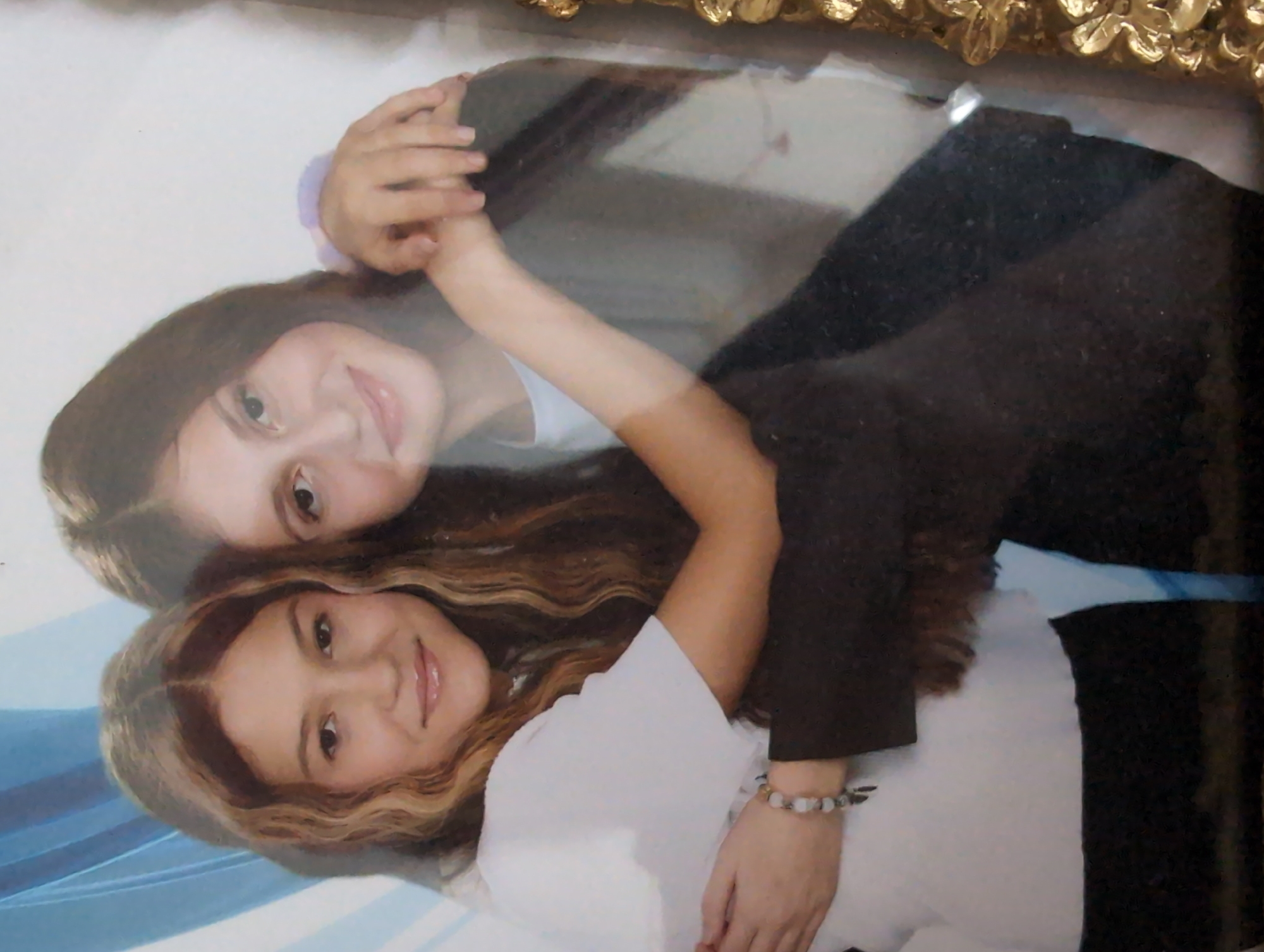 The image, rotated 90 degrees counter-clockwise, displays a portrait of two young females embracing, likely indoors. The woman positioned in the foreground has wavy, light brown hair and is wearing a white short-sleeved top. She smiles gently towards the viewer and wears a light-colored beaded bracelet on her left wrist. Behind her, a second woman with dark, straight hair and a light-colored top (possibly light blue or lavender) is embracing her. Her arms are wrapped around the foreground woman's shoulders, with their hands clasped near the foreground woman's right shoulder. Both subjects are smiling warmly, conveying an affectionate and happy mood. The background behind them features a soft, abstract blend of light blue and white. The entire image exhibits significant glare and reflections, particularly across the center and upper parts, indicating it is a photograph taken of a printed picture, possibly behind glass in a frame. A portion of a decorative, golden ornate frame is visible along the top edge of the overall image. No text is visible.