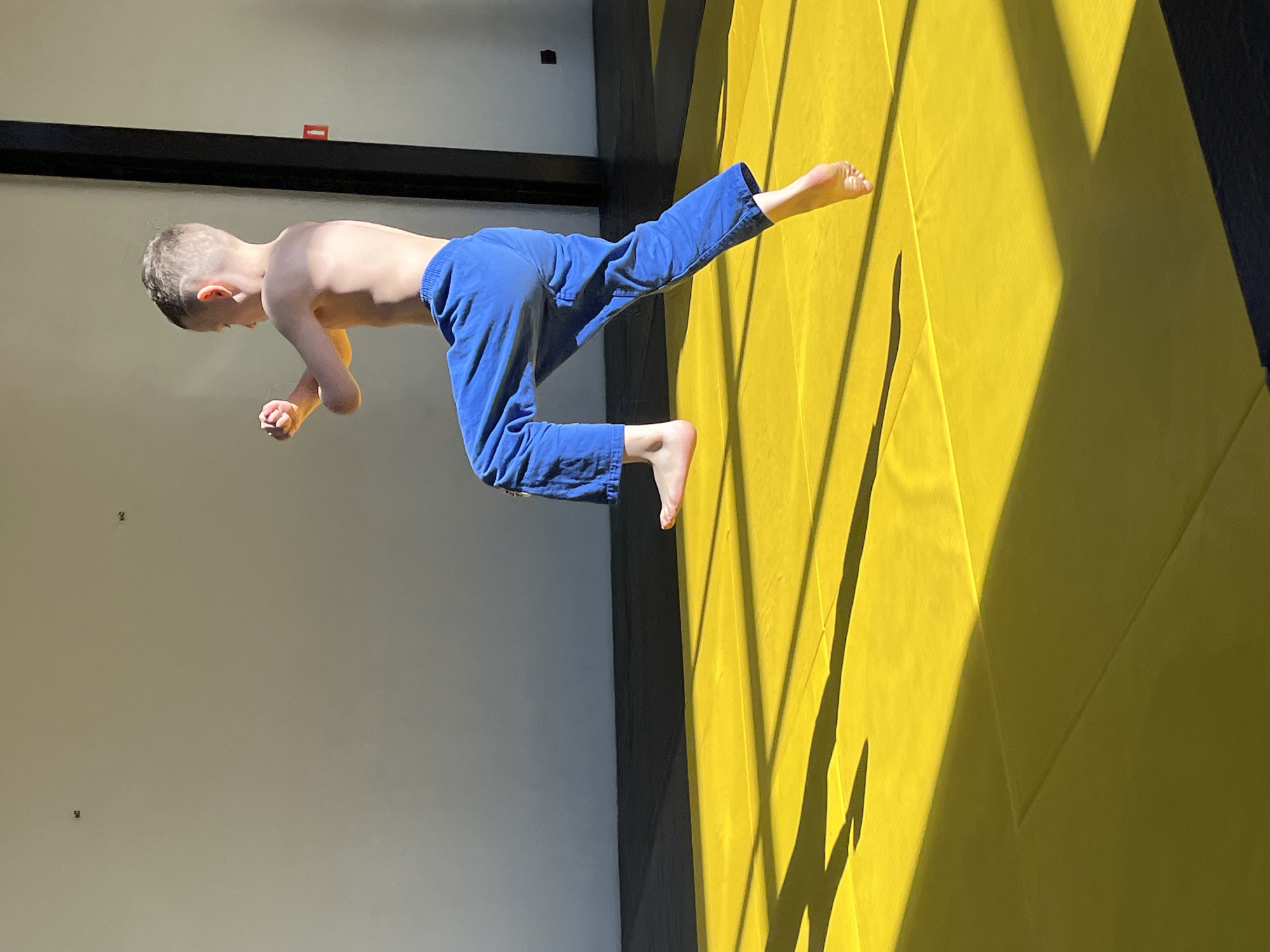 A young child wearing blue pants is positioned horizontally in the center of the frame. The child's upper body is bare. The child is indoors, on a yellow mat in a building in Krasnoyarsk, Russia. The background consists of a light-colored wall on the left and dark structural elements overhead. Shadows are cast on the yellow mat.