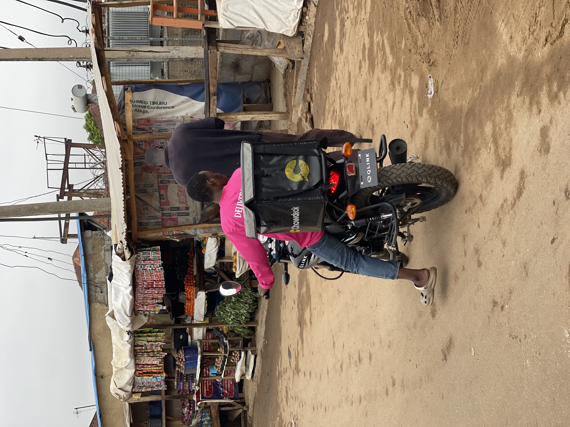 A motorcycle, bearing a "Chowdeck" delivery bag and a license plate reading "QLINK", is parked on a street in Kuchingoro, Nigeria. A delivery person in a pink shirt is on the bike with another person. The scene is adjacent to a roadside market stall, stocked with produce and packaged goods. Overhead, electrical wires crisscross the sky. Nearby is a sign mentioning "AHMED TINUBU" and a National Conference in Abuja.
