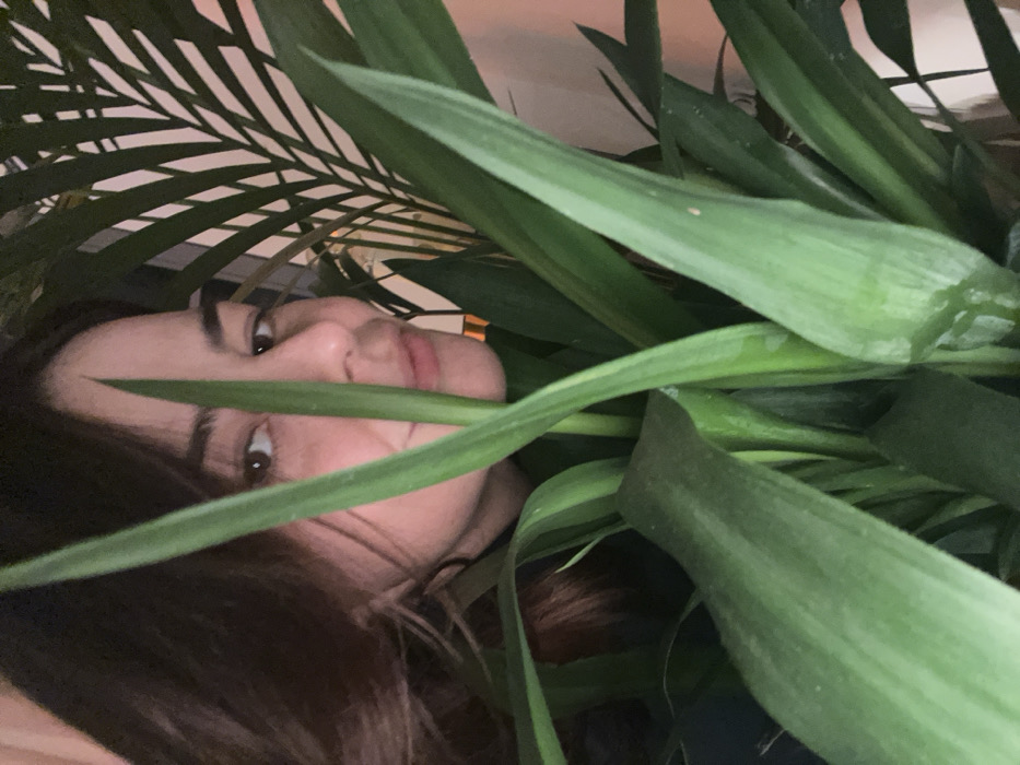 A person peeks through the dense foliage of several houseplants. The individual's face, with dark hair, is angled slightly, gazing out from behind and between the long, green leaves of a yucca-like plant on the right and the feathery fronds of a palm on the left. The lighting casts shadows, creating a playful and slightly obscured portrait. The person's expression is soft and curious, enhancing the intimate and whimsical feel of the image.