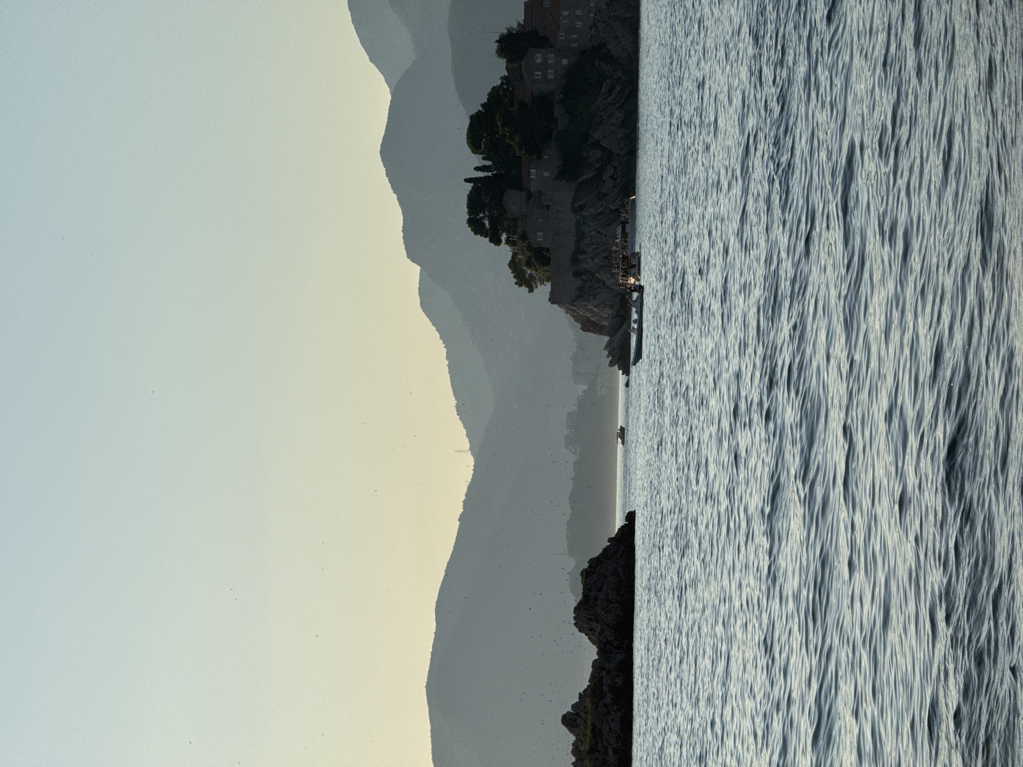 This image captures a serene coastal scene in Sveti Stefan, Montenegro, likely during the late afternoon or early evening, given the soft, diffused light. The primary subjects are the dramatic silhouettes of layered mountains receding into the distance, creating a sense of depth and scale against a pale, hazy sky.
Dominating the right side of the frame is the textured surface of the Adriatic Sea, its waves rendered in shades of grey and white, suggesting a gentle breeze. A rocky islet, characteristic of Sveti Stefan, juts out from the water on the right, topped with lush green trees and ancient stone buildings. The architecture, with its visible windows and tiled roofs, hints at a historical settlement.
On the water, a small boat is visible near the islet, its presence subtle against the vastness of the sea and mountains. The overall mood is tranquil and picturesque, evoking a sense of peace and timelessness. The faint scattering of birds in the sky adds a touch of natural dynamism to the otherwise still landscape. No visible text is present in the image.