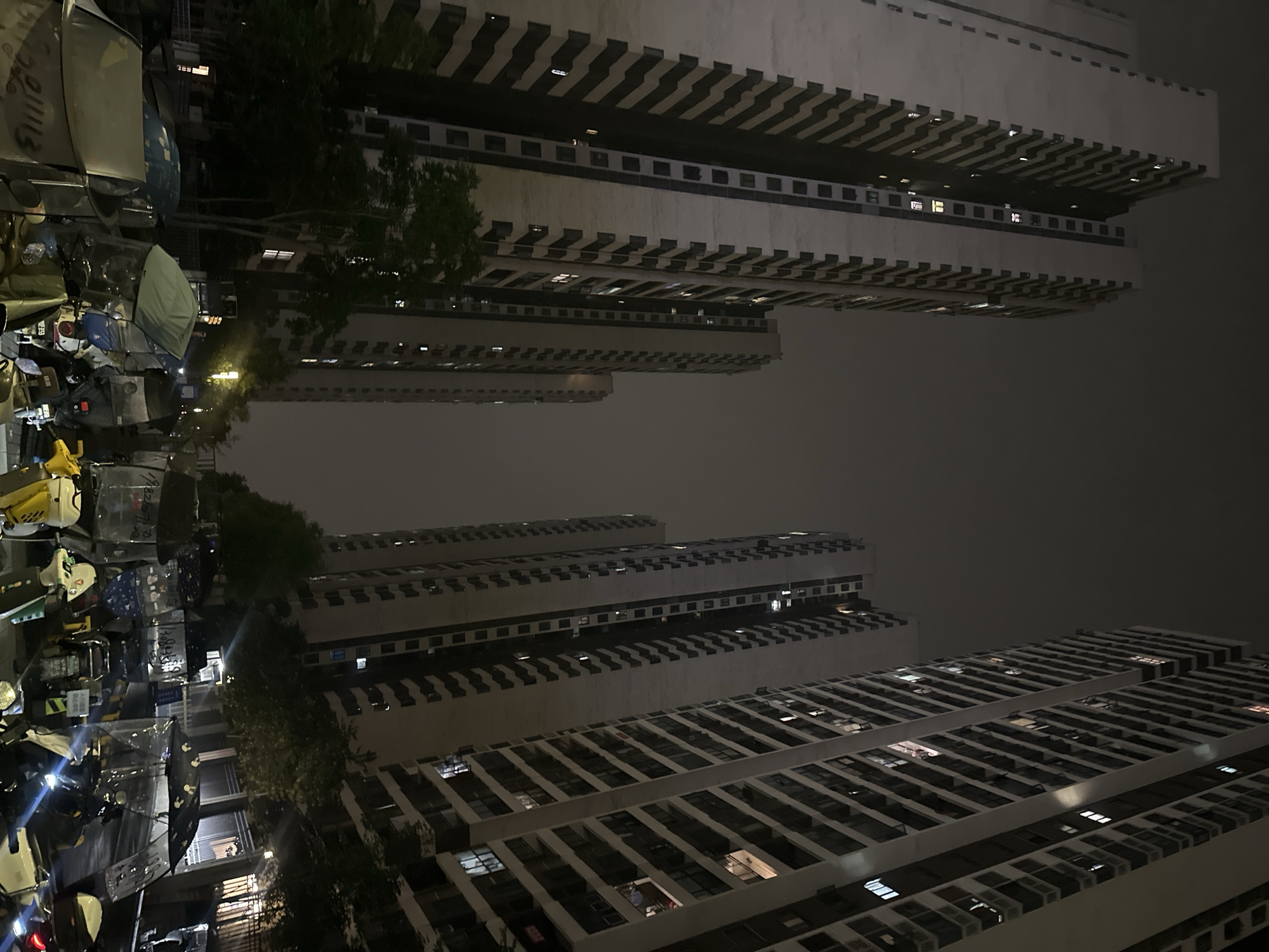 The image depicts a nighttime scene in Chongqing High-tech Zone, China. The composition is vertically oriented and tilted, featuring tall, densely packed apartment buildings receding into the distance under a dark, overcast sky. The buildings are characterized by repetitive horizontal and vertical lines formed by windows and balconies, some with visible interior lights. In the foreground, a collection of motorcycles and scooters is visible along what appears to be a street or parking area. Some of the vehicles are covered with tarps or protective coverings. A few trees with dark foliage are interspersed between the buildings and vehicles. The overall lighting is dim, contributing to a subdued and urban atmosphere.