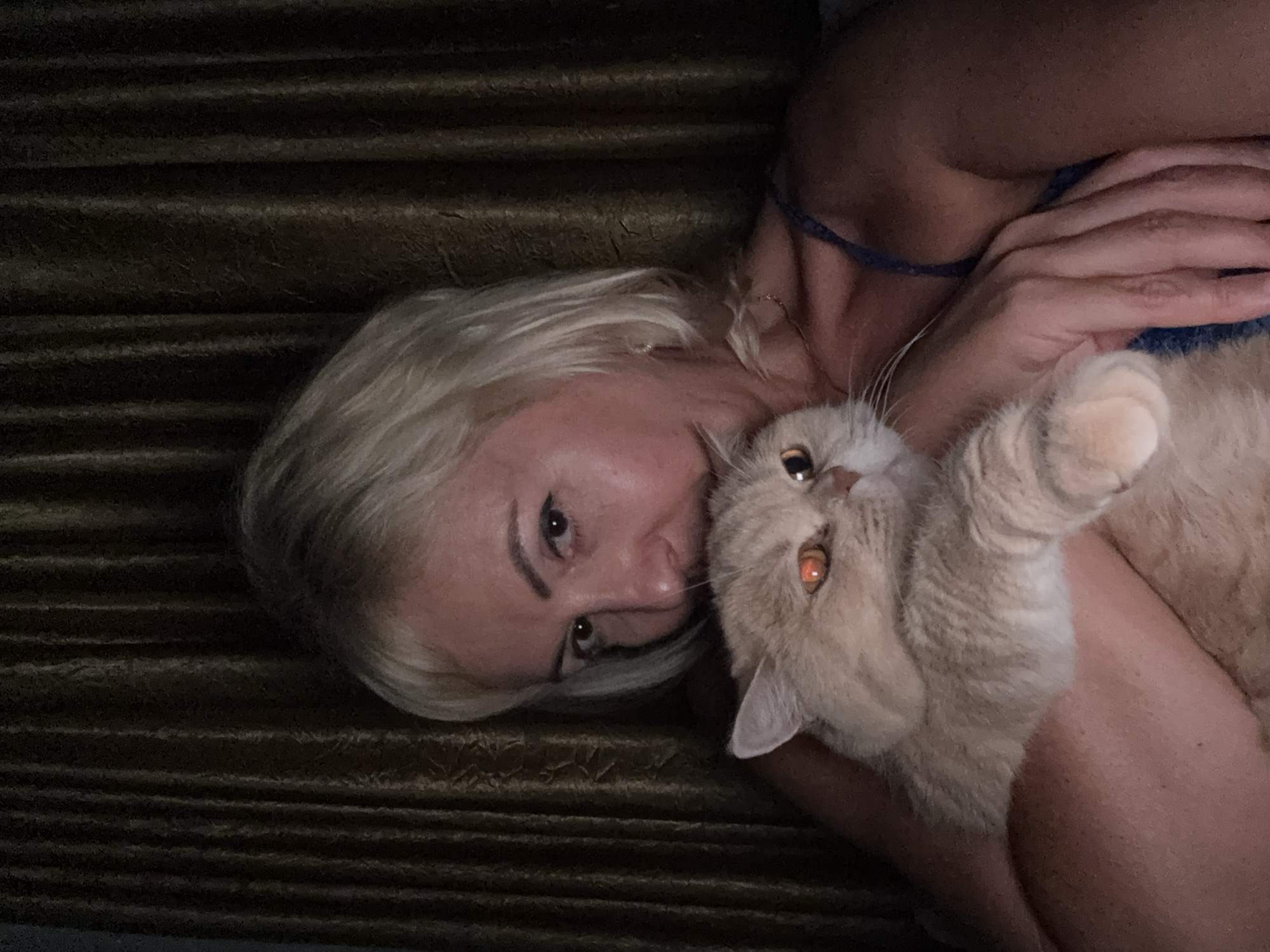 An outdoor, low-angle, medium shot shows a woman and a cat indoors in front of a dark, vertically pleated curtain. The woman has short blonde hair and is wearing a dark blue strap. She is holding a light-colored cat. The cat's eyes are reflecting orange light. The woman's left arm is wrapped around the cat, and her right hand is resting on the cat's body. The woman is looking at the camera. The lighting suggests it is nighttime, and the scene appears to be within a private residence, possibly a bedroom or living room. There is no indication of the city of Alanya, Turkey, in the image.