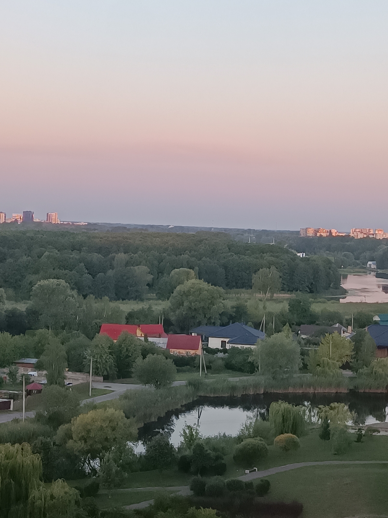 This is a high-angle, full-shot photograph of a serene landscape at dusk. The sky is a soft gradient of pale blue to pink, indicating the end of the day. Below the sky, a vast expanse of lush green forest dominates the middle ground. Scattered within the forest are several residential buildings, some with red roofs, suggesting a suburban or village setting.
In the foreground, a calm body of water, likely a lake or river, reflects the muted colors of the sky and the surrounding greenery. The water's edge is lined with tall reeds and various types of trees, including weeping willows. A paved path meanders through a well-maintained grassy area in the very foreground. In the distance, several modern high-rise buildings are visible on the horizon, silhouetted against the fading light, contrasting with the natural beauty of the foreground and middle ground.
The overall mood is peaceful and tranquil, with no visible people or animals. The lighting suggests it is either sunrise or sunset, with the soft, diffused light characteristic of these times. The scene captures a blend of nature and human habitation, typical of the outskirts of a city like Brest, Belarus. There is no visible text in the image.