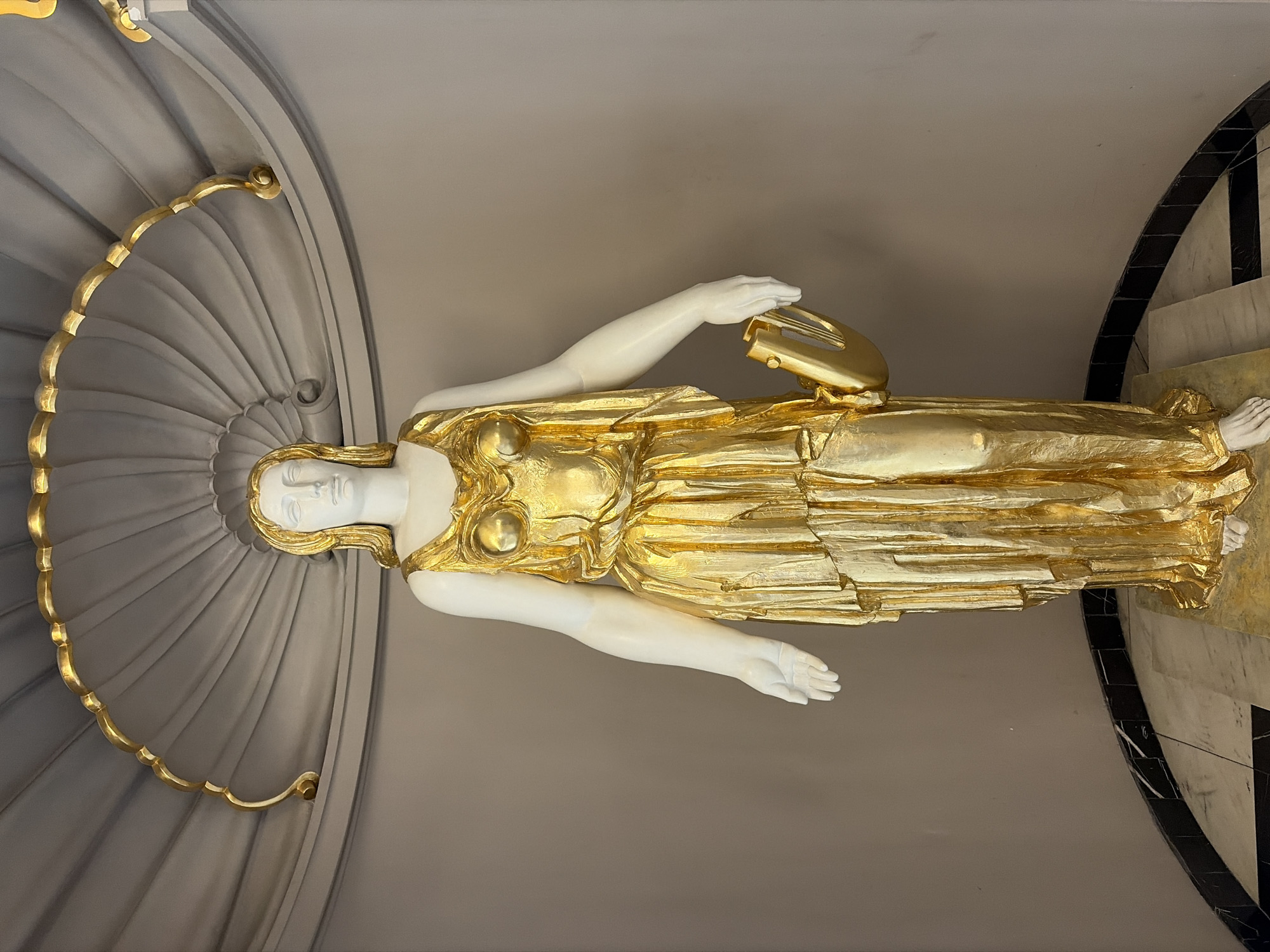 This media file features a life-sized statue of a woman in a gilded robe, holding a lyre. The statue is depicted in a vertical orientation, with the woman's head and upper body on the left side of the frame and her legs extending towards the right. She has a serene expression, with her eyes closed and her head tilted slightly. Her body is adorned with a gold-colored garment that appears to be draped and pleated, creating a sense of movement and flow. The lyre she holds is also gold and has a classical design.
The statue is positioned against a backdrop of a grey, fluted architectural element that resembles a fan or a shell. This element is further enhanced by a decorative gold trim along its curved edge. To the right of the statue, a portion of a black and white checkered pattern is visible, suggesting a floor or a decorative wall feature.
The lighting in the image is soft and even, casting a gentle glow on the golden parts of the statue and highlighting the smooth texture of the white marble-like material used for her face and arms. The overall impression is one of elegance and timeless beauty, evoking a sense of classical art and mythology. The environment appears to be indoors, possibly in a museum, gallery, or a grand architectural space. The location context provided is Sofia, Bulgaria, suggesting the statue might be a significant artwork within the city. There is no visible text in the image.