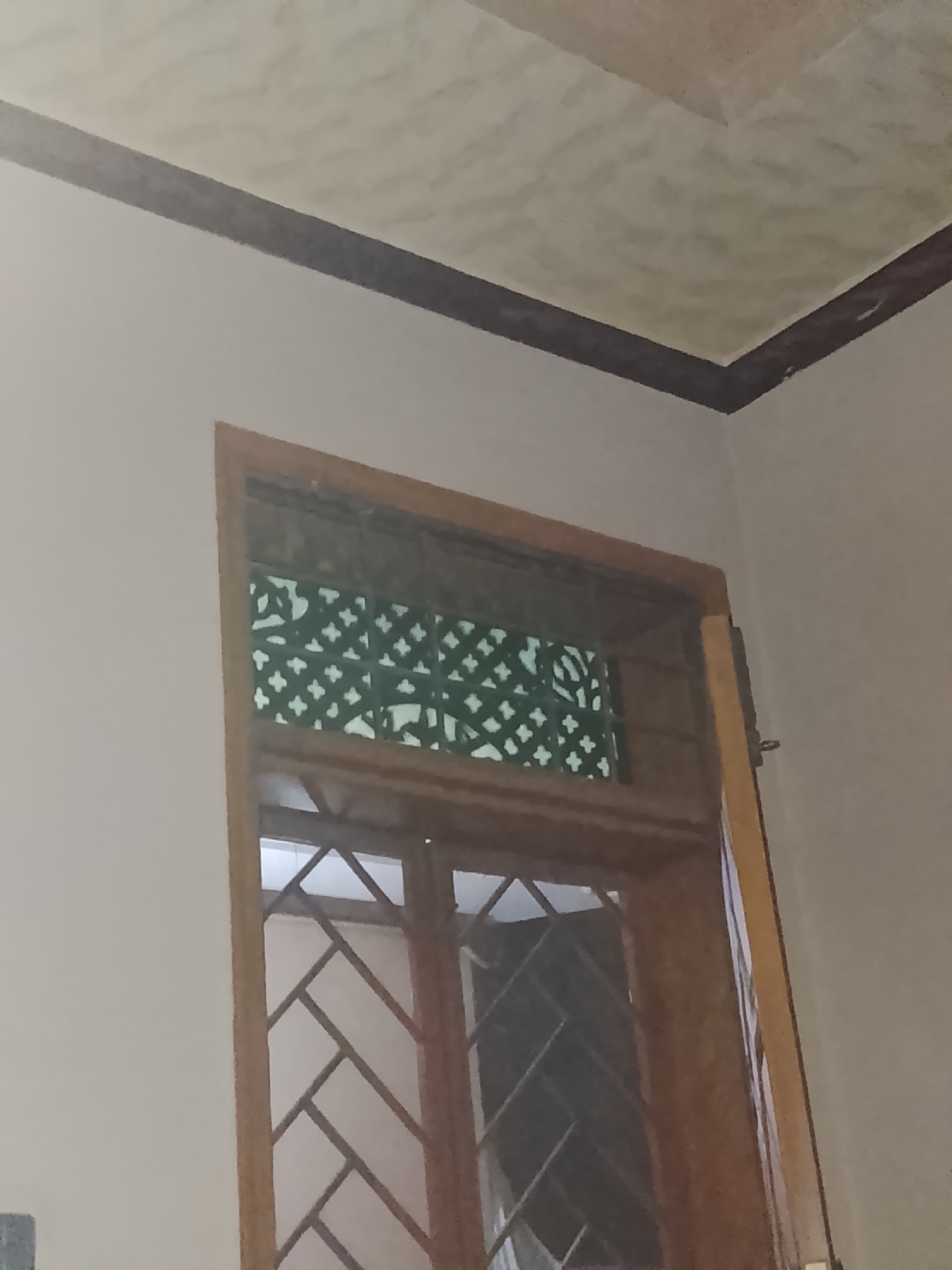 The image displays an interior architectural detail, specifically focusing on a wooden window or door frame against a light-colored wall. The frame is made of polished or stained wood, showcasing a rich, warm brown hue. Above the main section of the window/door, there is a transom or vent area featuring a decorative green-colored grating with a repeating geometric criss-cross pattern, resembling a jali. The lower part of the window/door also has a wooden grid pattern, possibly for glass panes or a screen, though the area behind the grid appears dark.
The wall is painted a plain, light beige or off-white color. A dark brown trim or border runs horizontally along the top of the wall, separating it from the ceiling, and then turns sharply at a corner, extending upwards on the adjacent wall. The ceiling is also light-colored, possibly off-white or light beige, and has a subtle, textured, or patterned finish that gives it a slightly mottled appearance.
No people are visible, and no specific activity or event is taking place, presenting a static interior scene. The lighting is indirect and appears somewhat dim, suggesting either ambient indoor lighting or diffused natural light from an unseen source. There is no visible text in the image. The setting appears to be an indoor space, possibly a home or building, with traditional architectural elements suggested by the decorative grating.