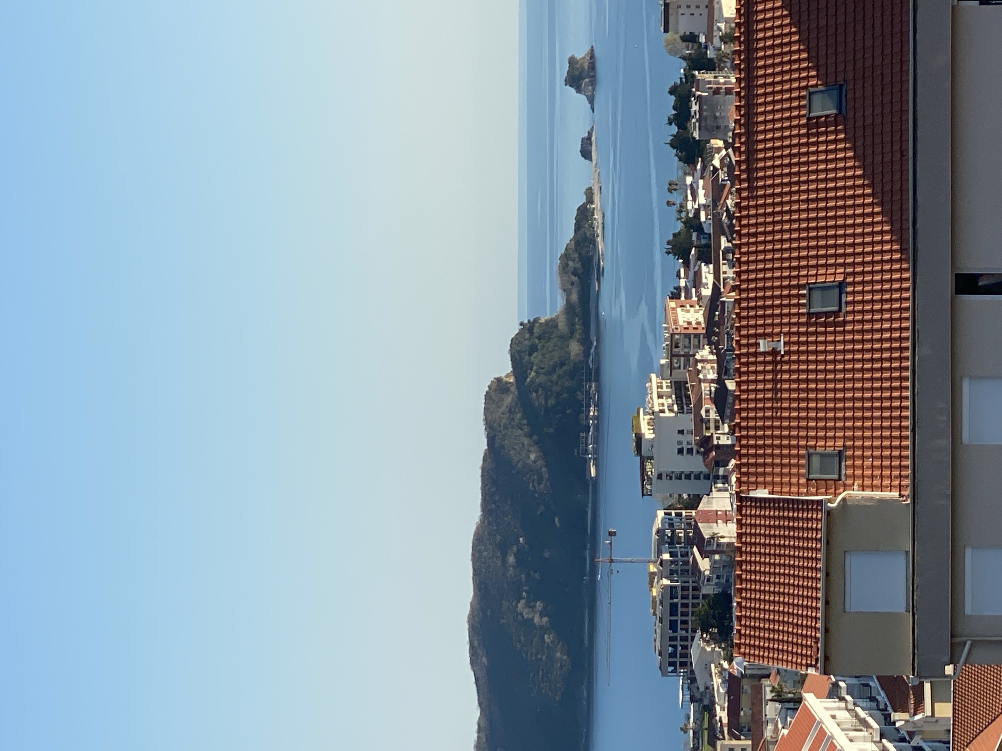 The image depicts a view from a high vantage point, likely a building or elevated ground in Budva, Montenegro. The scene is dominated by the Adriatic Sea in the background, with a small island or rocky outcrop visible in the distance. A densely forested hill runs along the coastline, separating the water from the town. The foreground shows a dense cluster of buildings, mostly residential, characterized by terracotta tile roofs and various architectural styles. The building where the picture was taken has its edge visible in the right of the image. The sky is clear and light blue. The lighting suggests a daytime scene.
