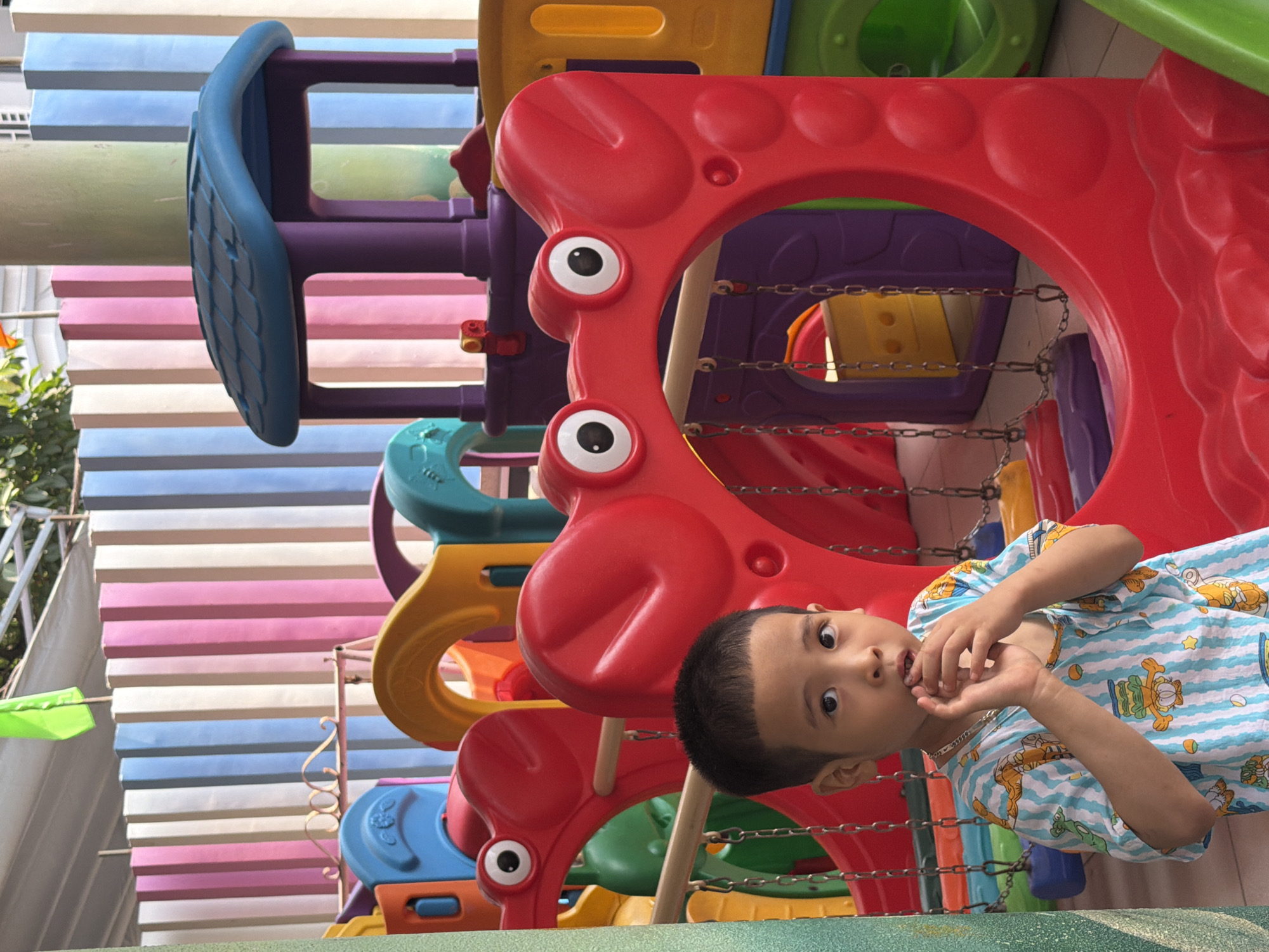 The image captures a child playing on a colorful playground structure. The playground features red elements with cartoonish eyes, a blue climbing structure, and various other colorful components. The child, wearing a light blue striped shirt with cartoon designs, is looking at the camera with a focused expression. The playground is located in Đà Nẵng, Vietnam. In the background, a wall with horizontal stripes of pink, white, and blue is visible.
