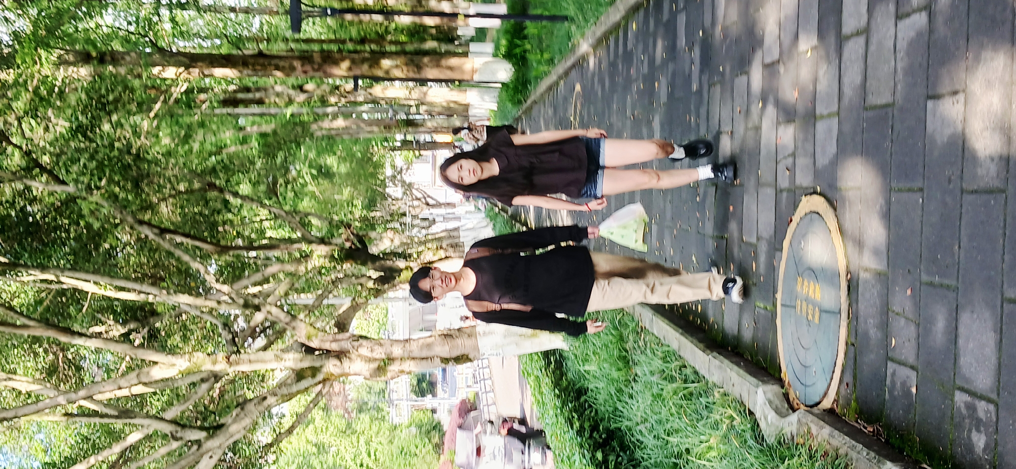 Two women walk on a brick path in Chongqing High-tech Zone, China. One woman wears a long-sleeved black shirt and khaki pants, with a backpack. The other wears a short-sleeved brown shirt and denim shorts. A plastic bag is held by the first woman, and a round manhole cover is nearby. Trees and grass line the path.