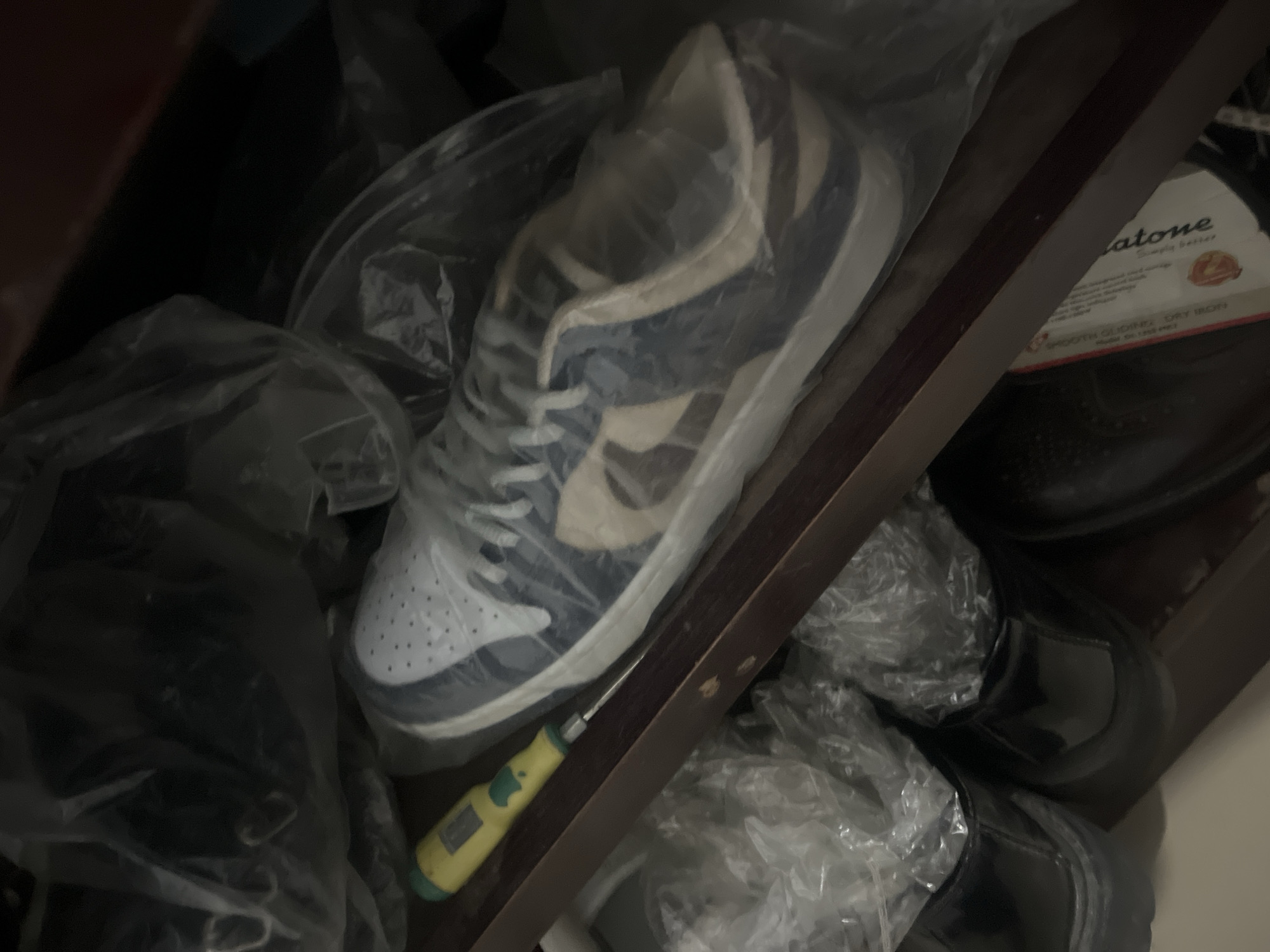 The image captures a dimly lit storage space, likely a shelf or cabinet, filled with various items, primarily shoes, stored within clear plastic bags. In the center, a pair of sneakers, possibly Nike Dunks, with white and blue-grey sections and white laces, is prominently displayed, enclosed in plastic. A small screwdriver with a bright yellow handle and a green end lies below and to the left of the sneakers.
To the right, partially obscured by a dark wooden divider, are black, formal-looking shoes, also appearing to be wrapped in plastic or stored with plastic. Further right, a white label attached to a brown object, possibly a box, is visible. The text on the label clearly reads "atone" followed by "Simply better." Below this, "SMOOTH GLIDING DRY IRON" and additional text, possibly "HIGH OR LOW HEAT," can be discerned, suggesting an iron or related product is also stored here. The scene depicts a static arrangement of stored belongings in what appears to be an indoor storage area in Abuja, Nigeria.